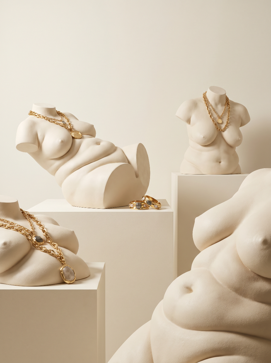 Sculptural art display featuring several white, marble-like torso sculptures adorned with gold jewelry, including necklaces and rings, arranged on white pedestals.