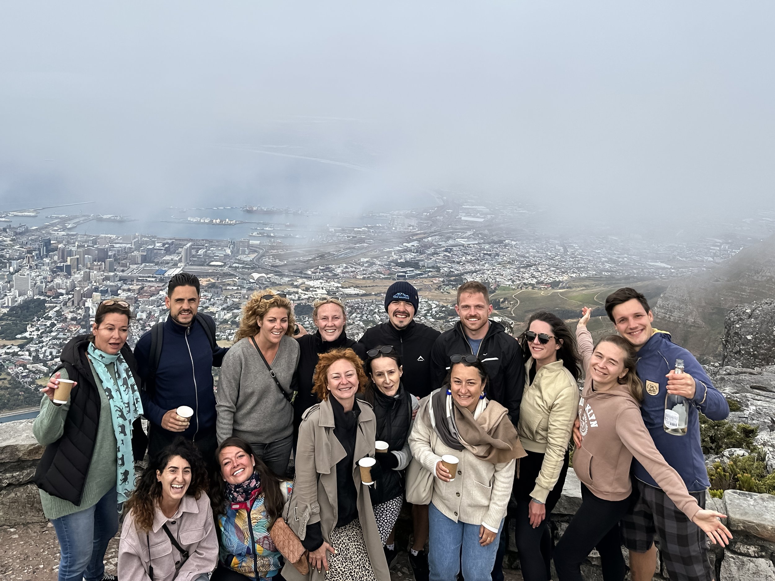 A group of digital nomads smiling and posing for a photo on a mountain with a city and water in the background, surrounded by fog.