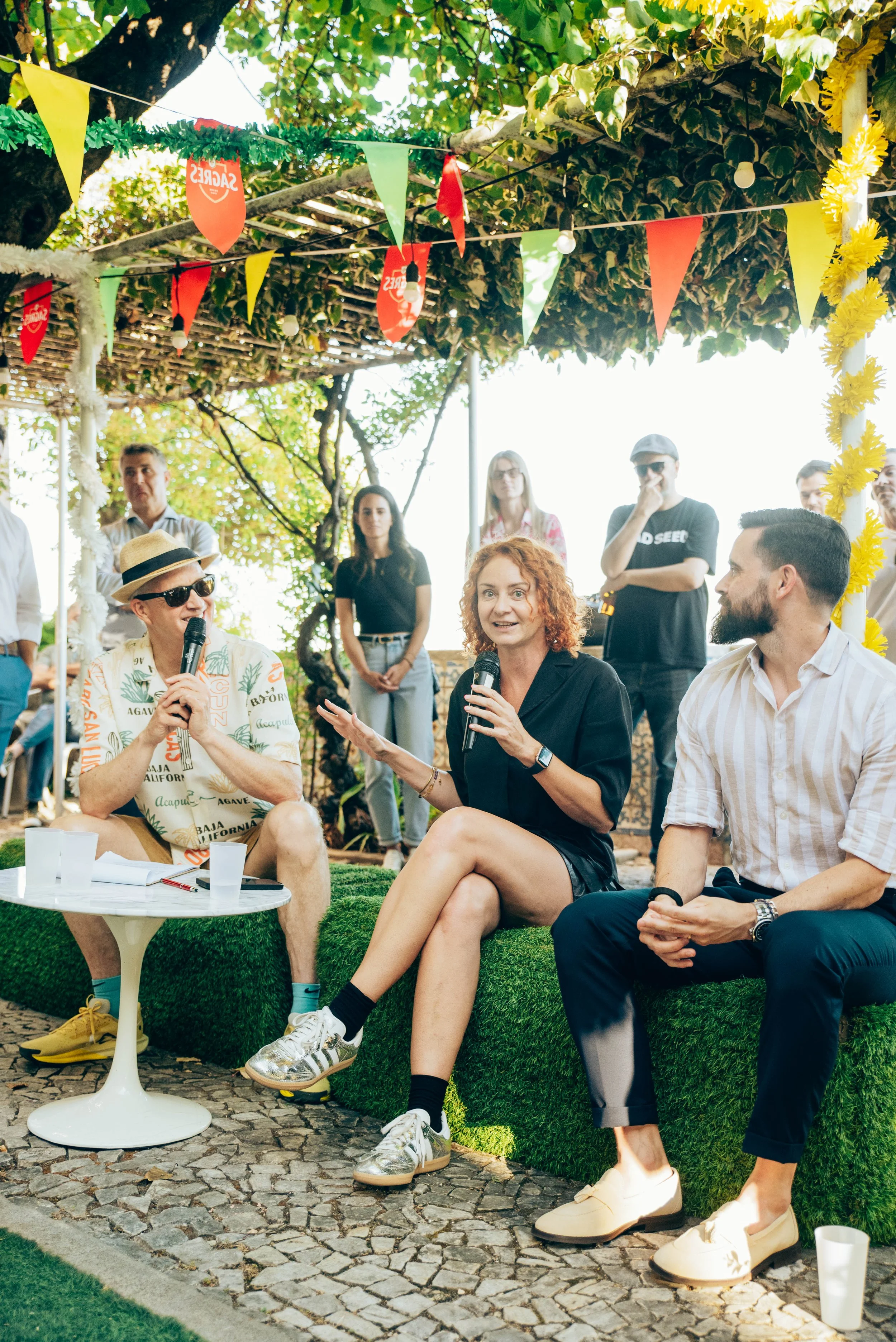 People gathered outdoors under a decorated canopy at a Red Bridge Lisbon event, engaged in a conversation or panel discussion, Kate speaking into a microphone and others listening, sitting on grass 