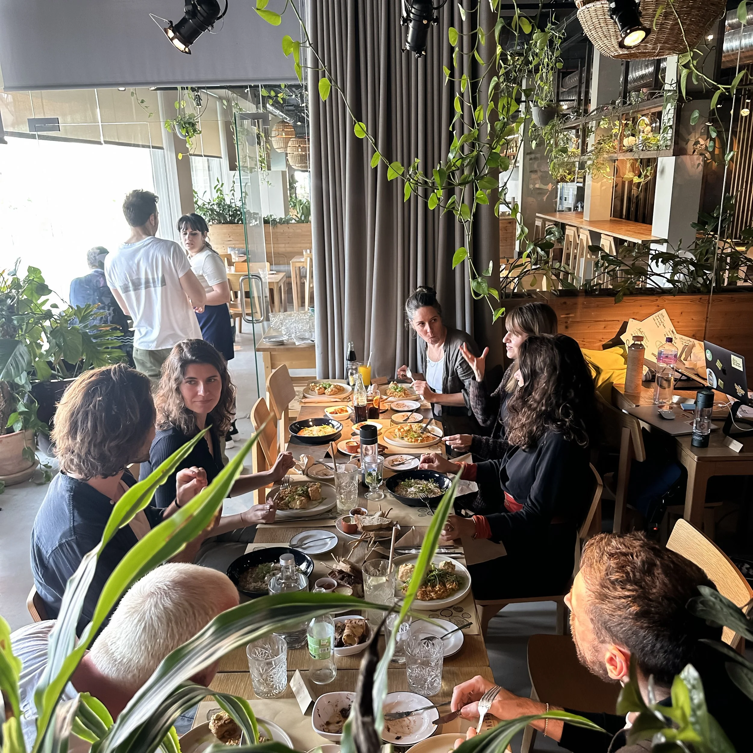 Coworking and Spa days in Ericeira, Portugal. People gathered around a long wooden table enjoying a meal in a restaurant filled with plants and natural light.