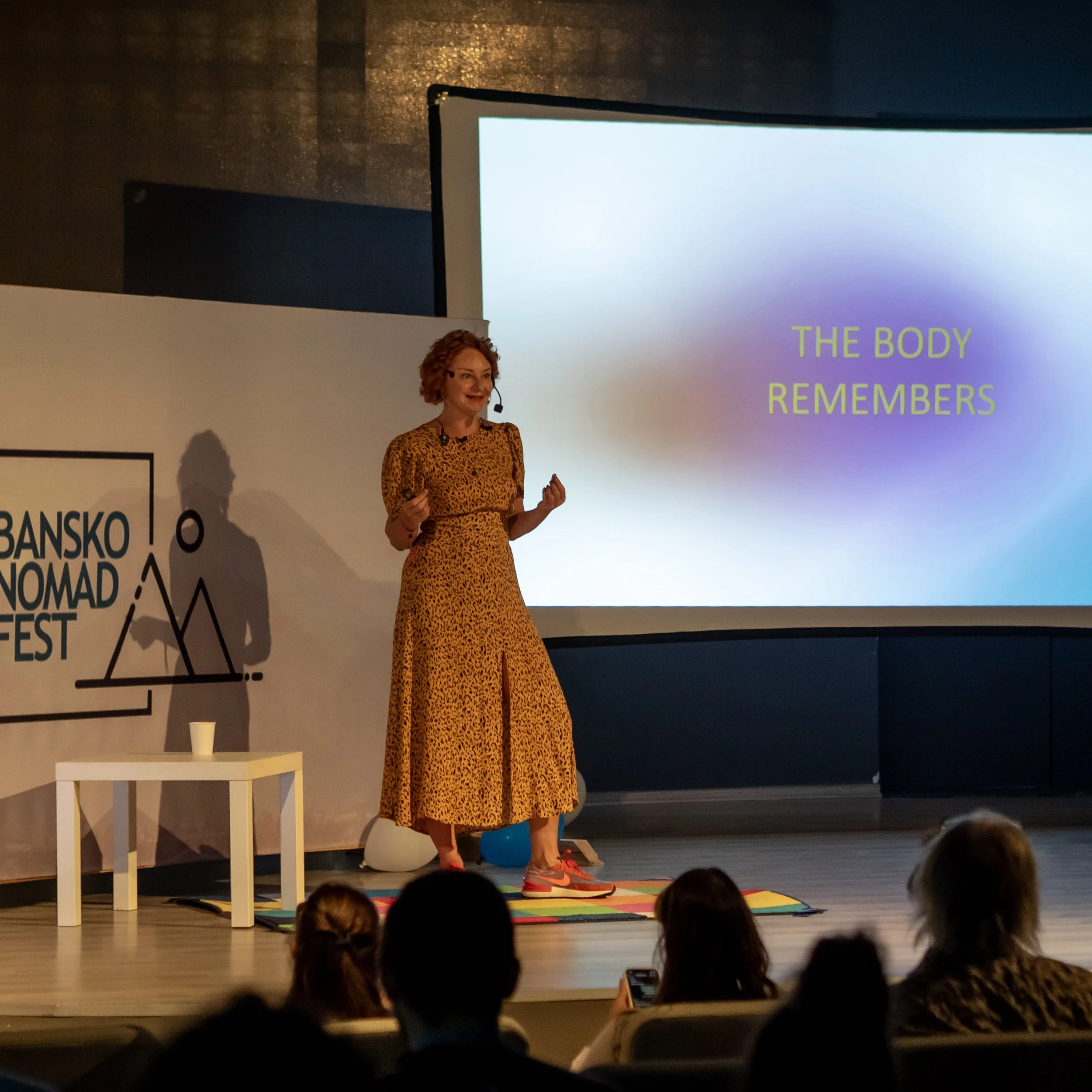  Kate Shifman is giving a presentation on stage at Bansko Nomad Fest in front of a large screen displaying the text 'THE BODY REMEMBERS.'  There is an audience in front of her, and a small table with a cup is nearby.