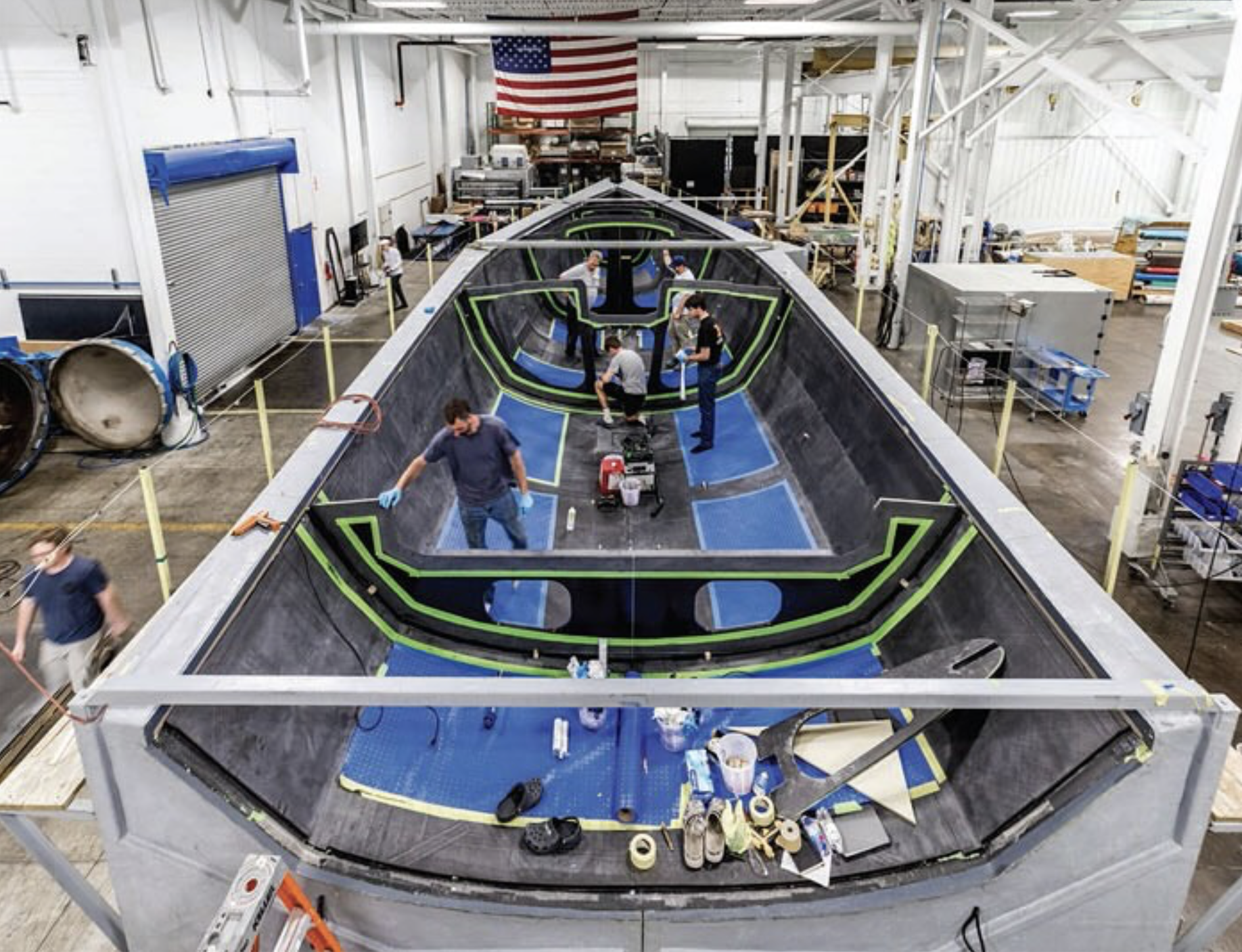 Employees working inside a manufacturing facility assembling a large spacecraft or aerospace vehicle fuselage. The structure is fed with materials, and the workers are installing and inspecting components around a central cockpit area. The American flag hangs above the workspace.