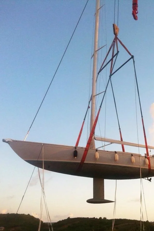 A sailboat lifted out of the water, hanging by straps, with the ocean and hills in the background.