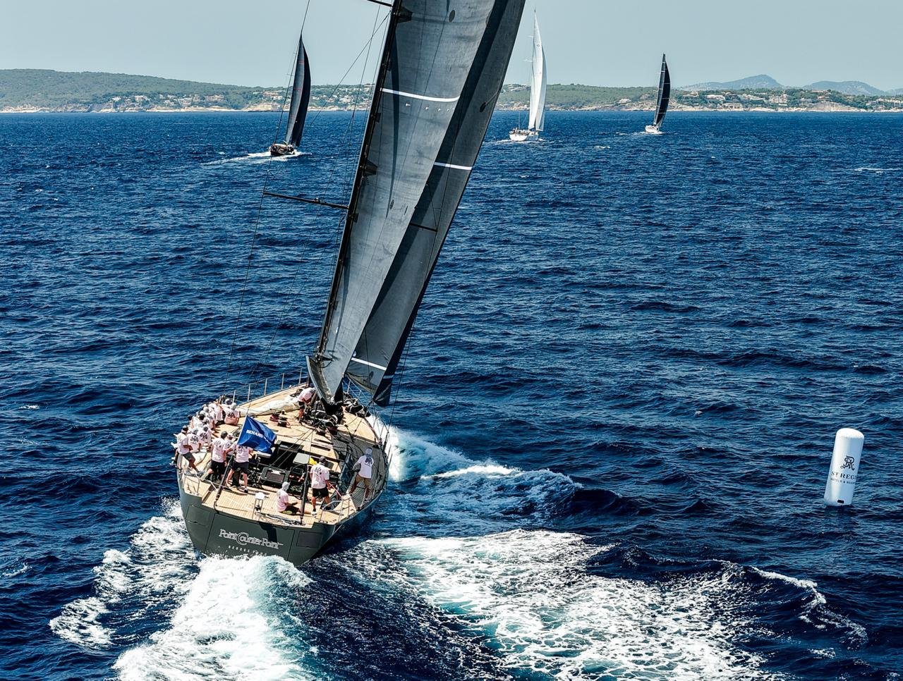 A black racing sailboat with multiple crew members on deck, sailing fast with black sails in open blue water, with other sailboats in the distance and a distant shoreline.