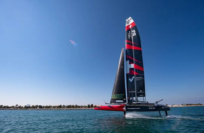 A high-performance racing sailboat with black, red, and white sails gliding on calm water under a clear blue sky.