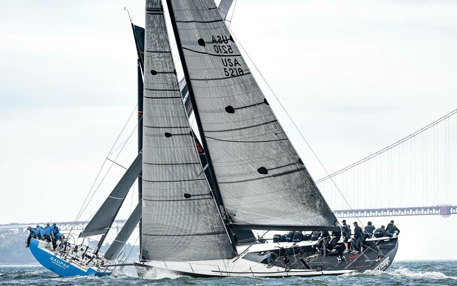 A large racing sailboat with black and gray sails, crew members on deck, and a city bridge in the background, moving through water.