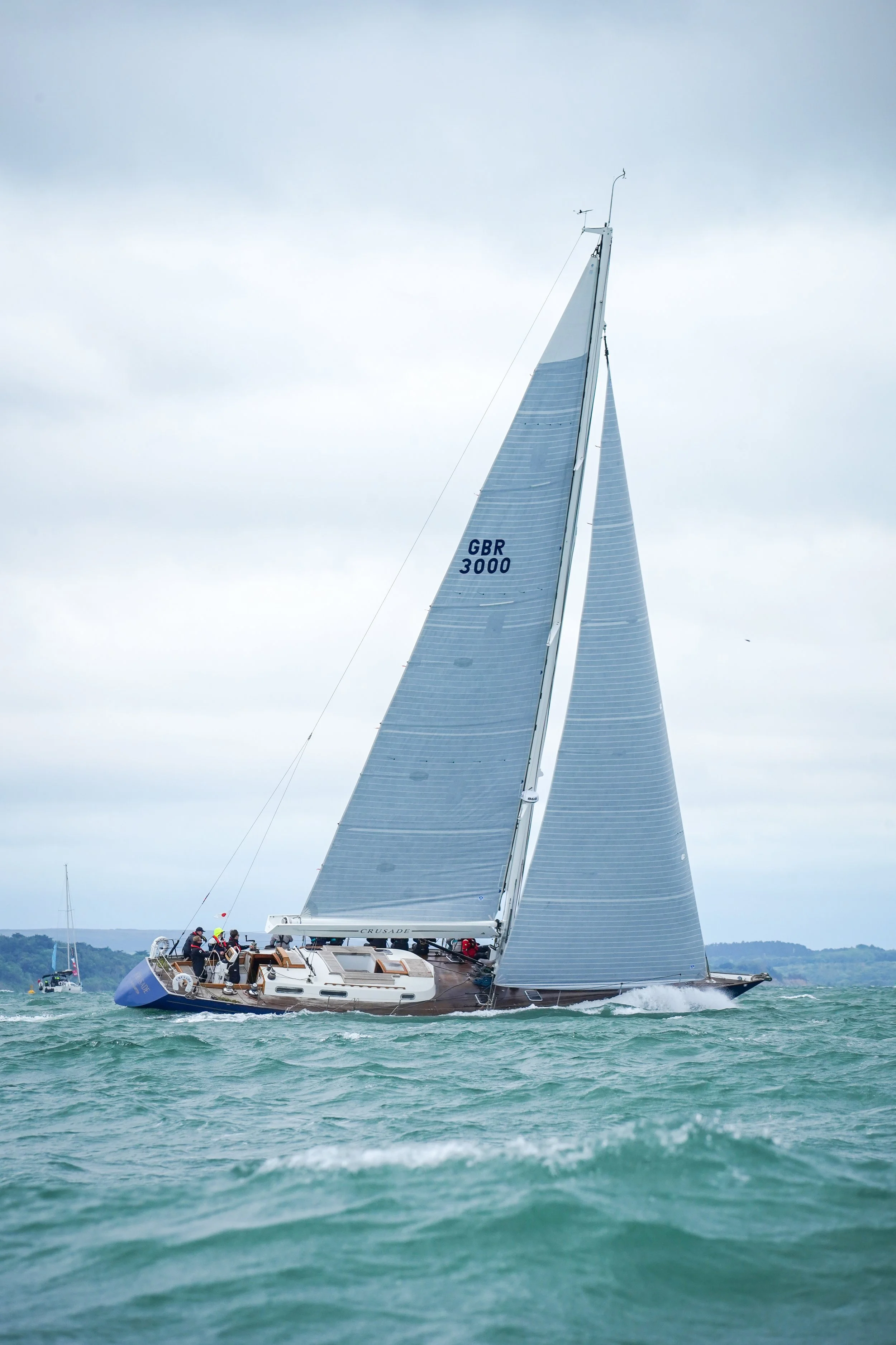 A sailboat with a crew sailing on the water, with gray sails marked 'GBR 3000' and a large main sail named 'CRUSADE.' Overcast sky with other boats visible in the background.