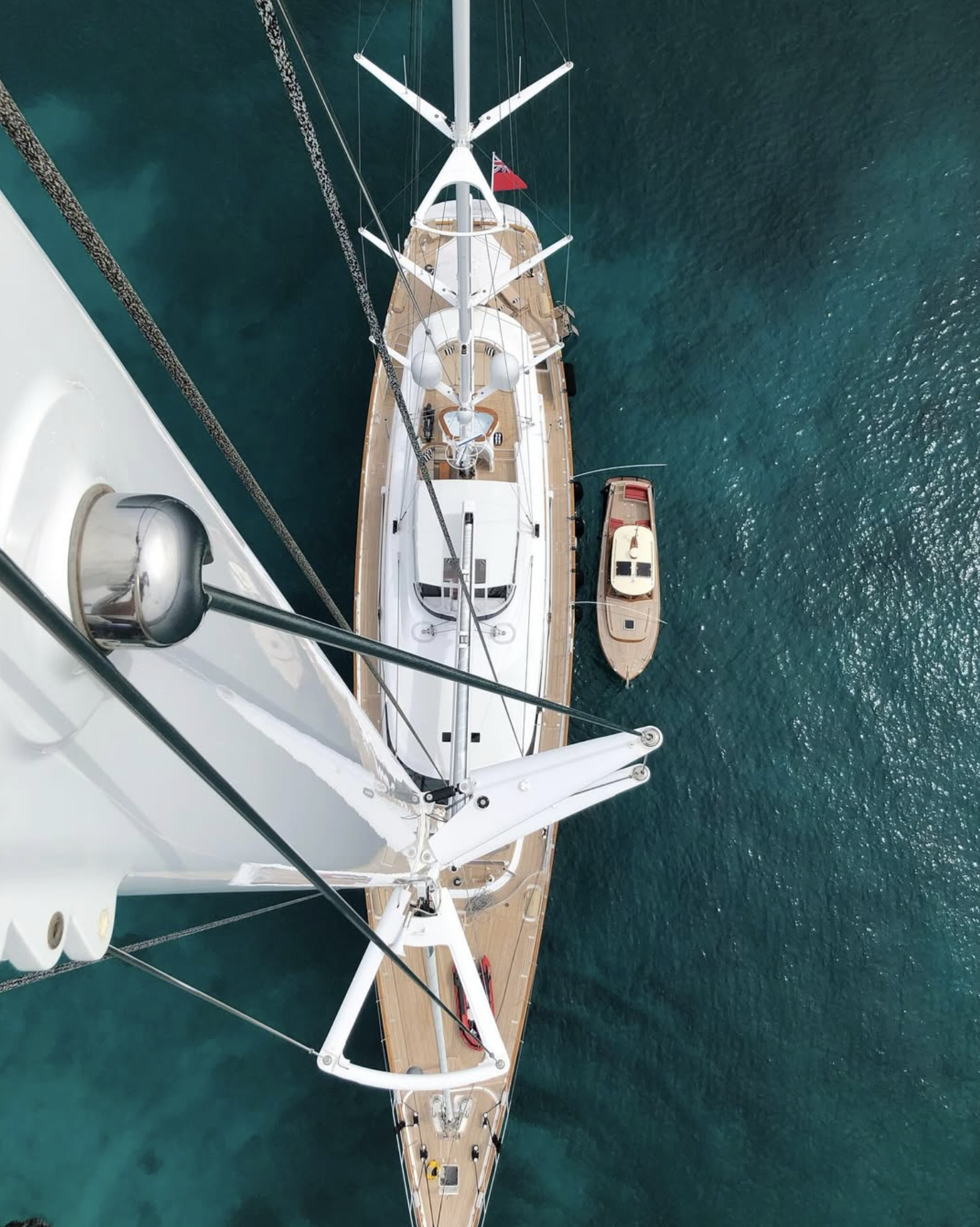 Aerial view of a sailboat and a smaller motorboat on calm blue water. The sailboat has a white mast and deck, with ropes and rigging visible, and a small red flag at the stern.