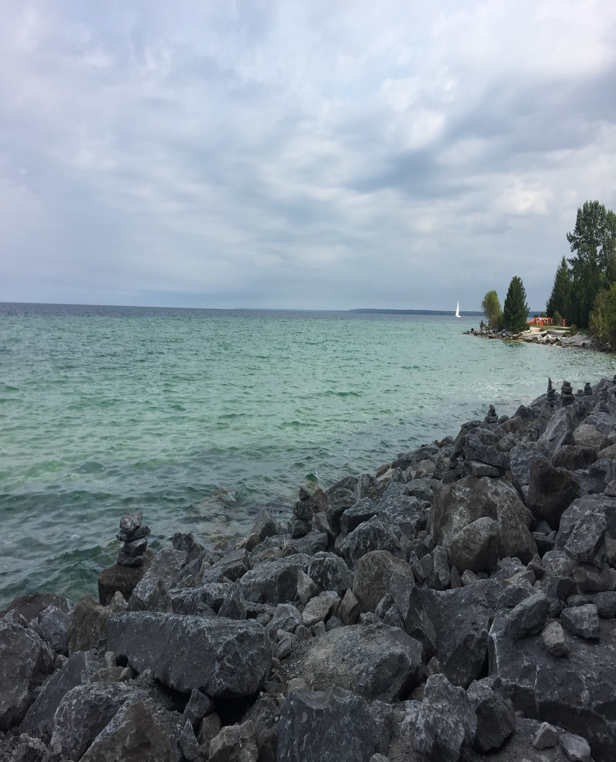 Lake with rocky shoreline, greenish water, trees along the right side, a small white sailboat in the distance, and a cloudy sky.