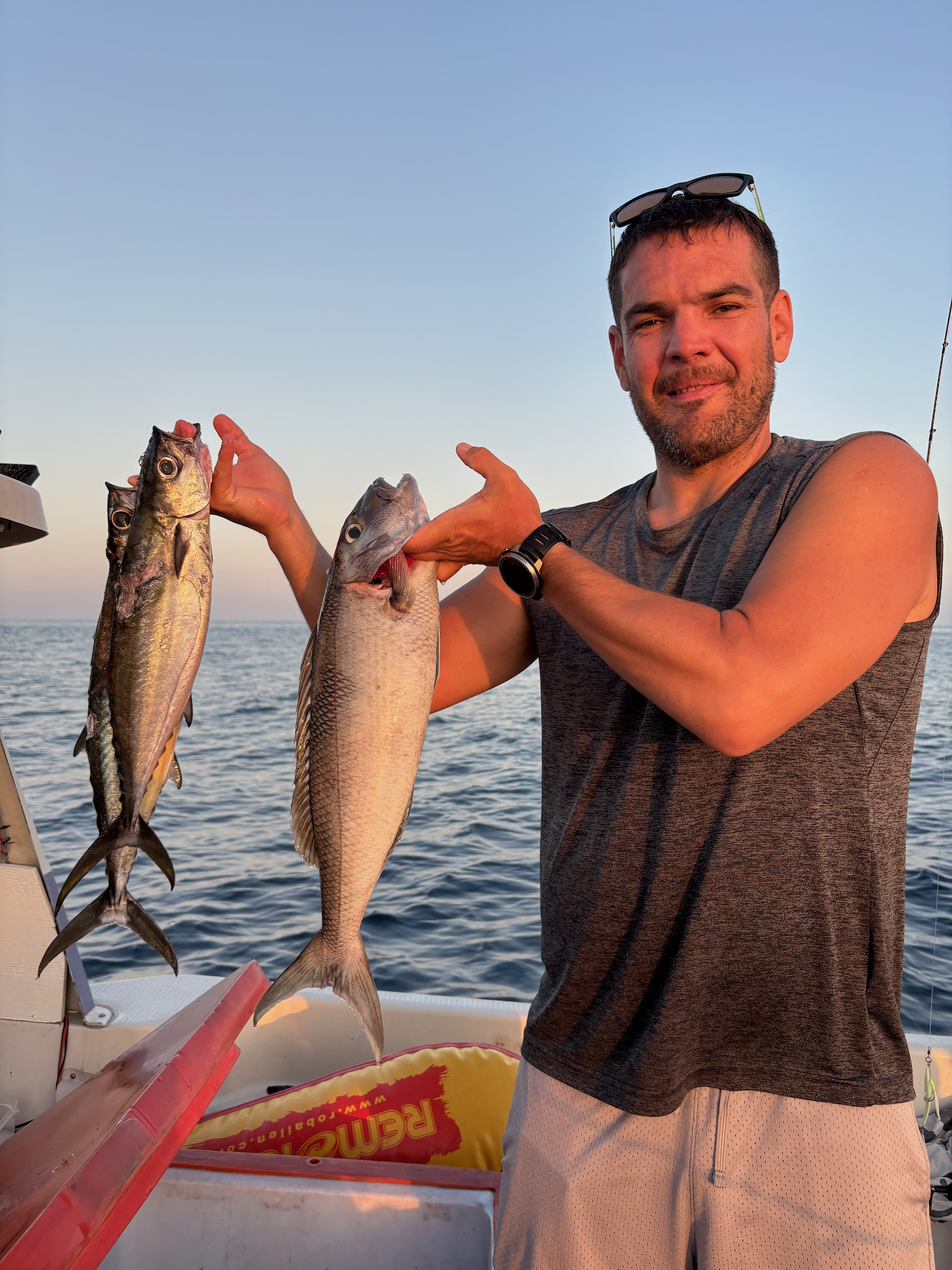 A man on a boat holding two fish, one in each hand, with the ocean and sky in the background during sunset.