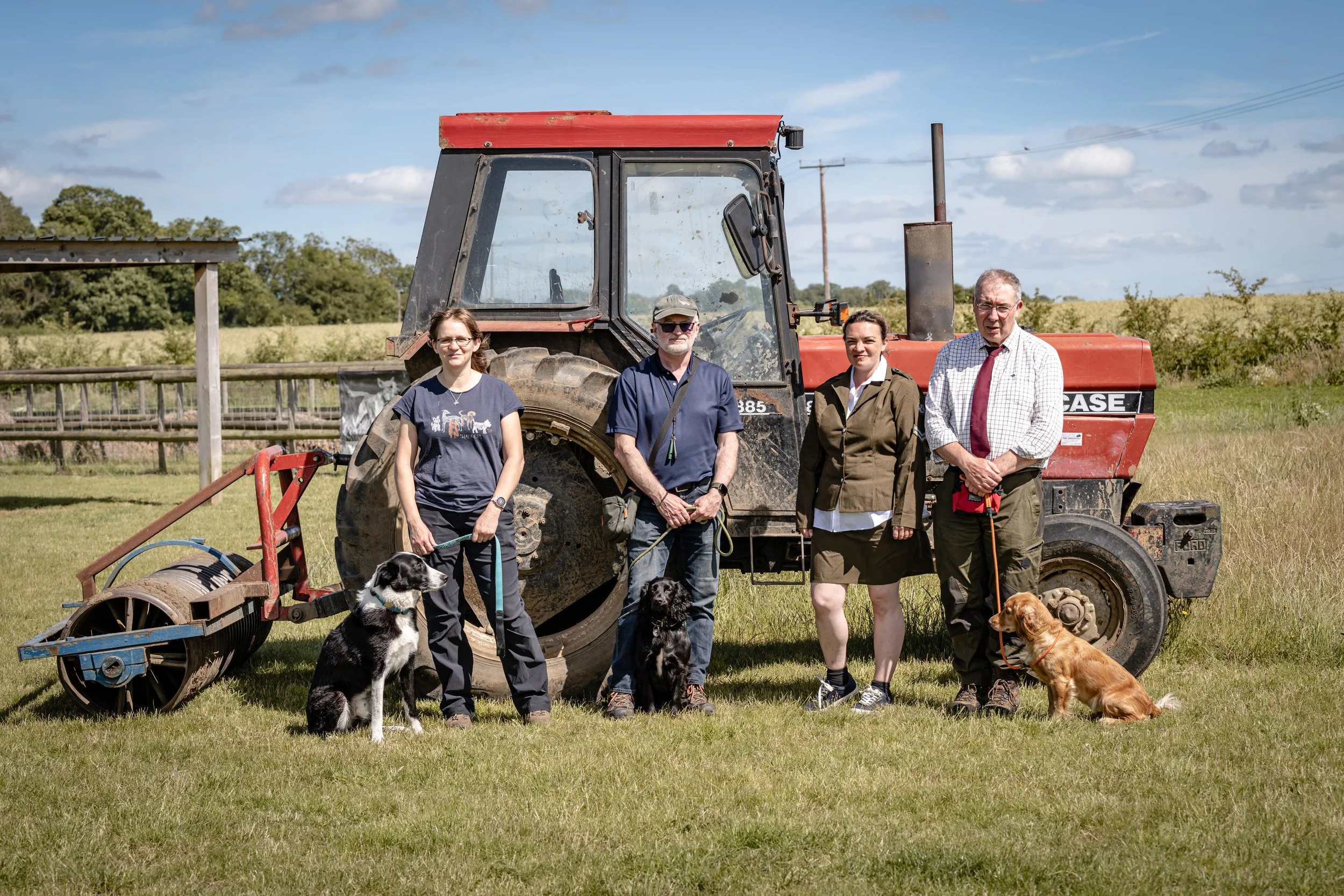 Four people standing in front of a red tractor with dogs on leashes in a grassy field, trees and power lines in the background on a sunny day.