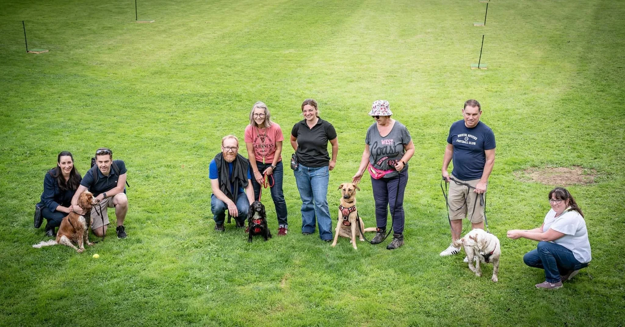 Group of adults and dogs on a grassy field, some sitting and some standing, with dog agility equipment in the background.