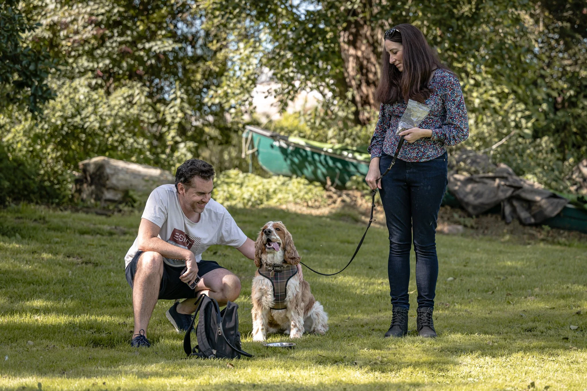 Two people and a dog outdoors on a grassy area with trees and a boat in the background. One person is kneeling, petting the dog, and the other person is standing, holding a leash.