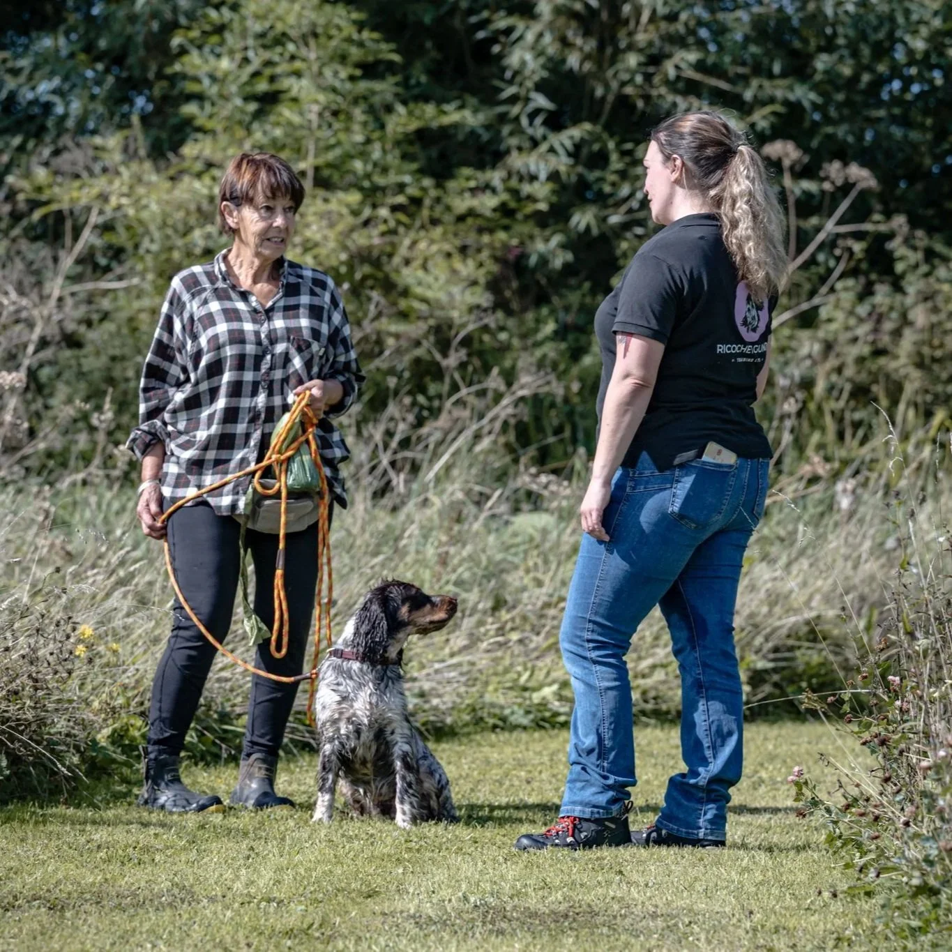 Two women having a conversation outdoors with a dog sitting on the grass between them, surrounded by trees and bushes