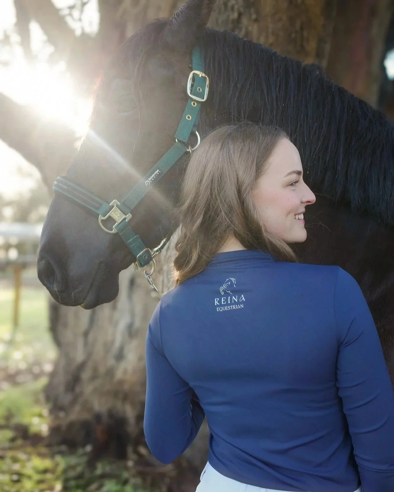 A young woman with long brown hair smiling and hugging a black horse outdoors during daytime, wearing a blue shirt with a logo and text that reads 'Reina Equestrian', with trees and sunlight in the background.