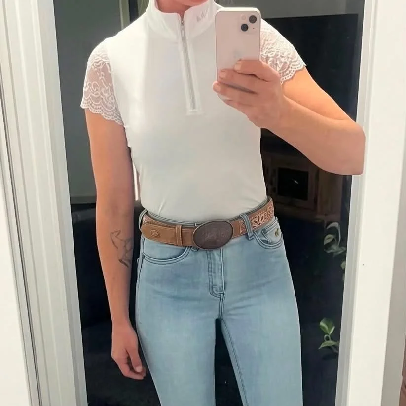 Person taking a mirror selfie, wearing a white top with lace sleeves, light blue jeans, and a brown belt with a large oval buckle. The person's face is not visible.
