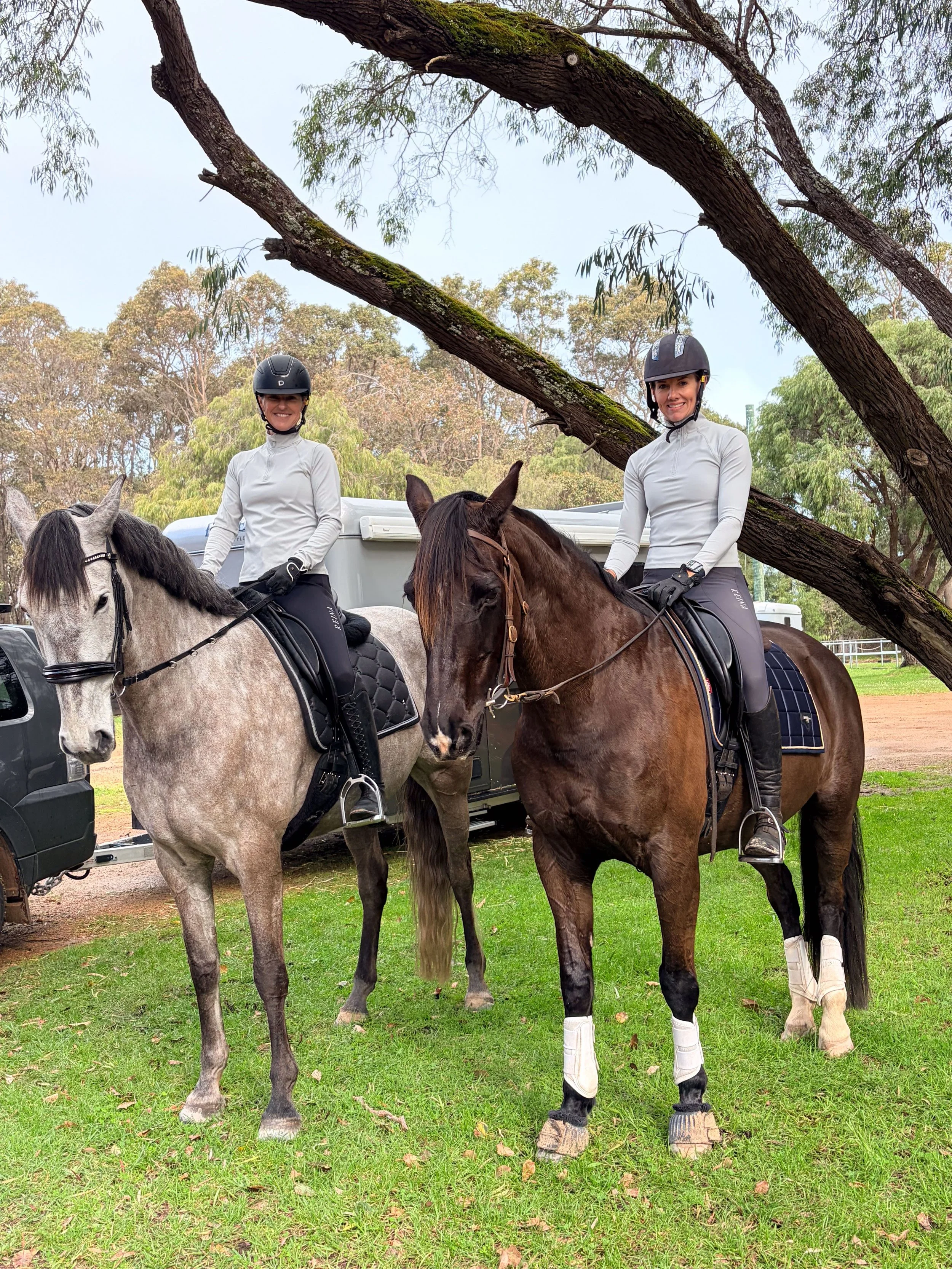 Two women riding horses outdoors in a park. They are wearing helmets and riding gear, sitting on brown and gray horses. There are trees and a trailer in the background.