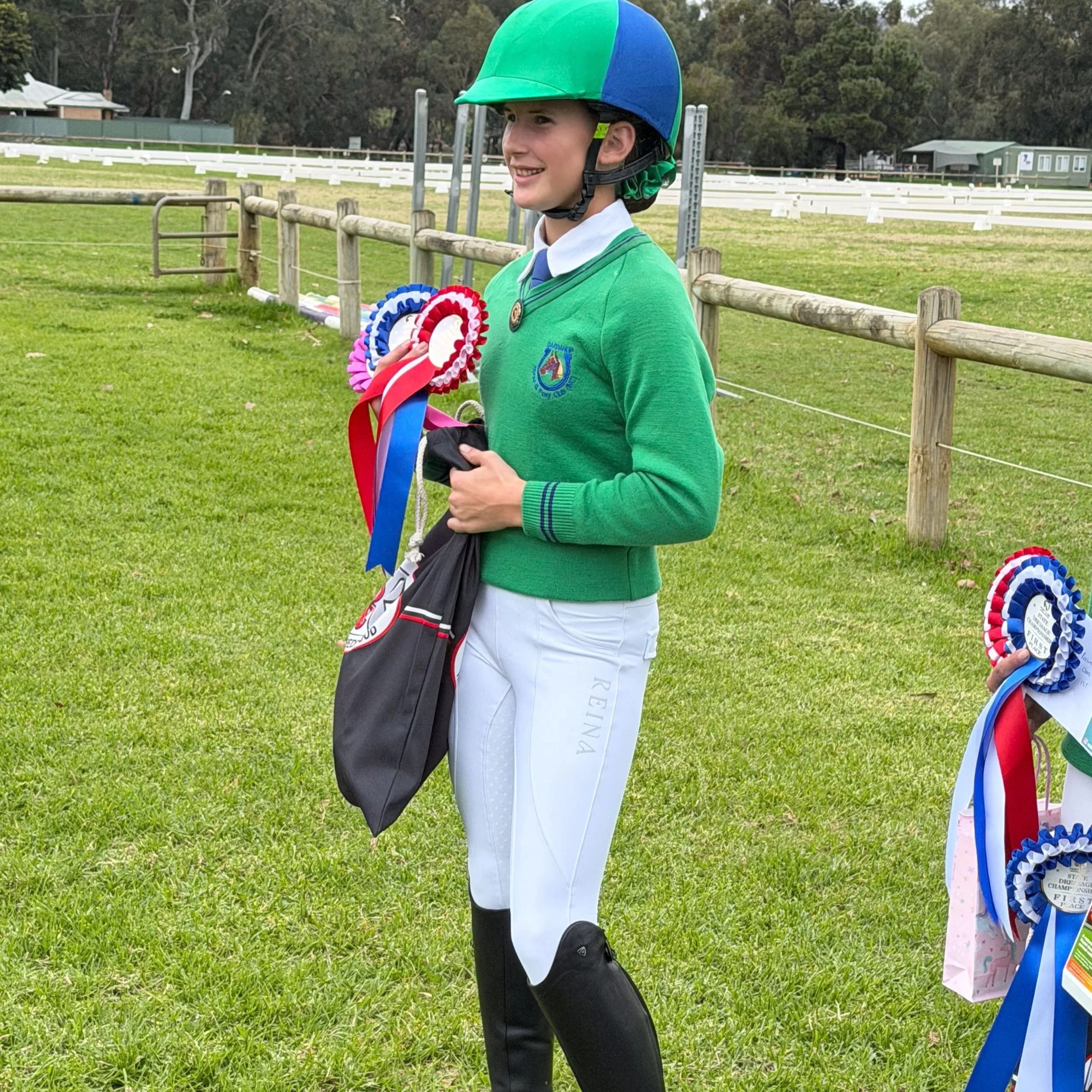 A young boy wearing a green jacket, white pants, and a helmet, holding colorful ribbons and standing next to a display of awards outdoors in a grassy area with a wooden fence and trees in the background.