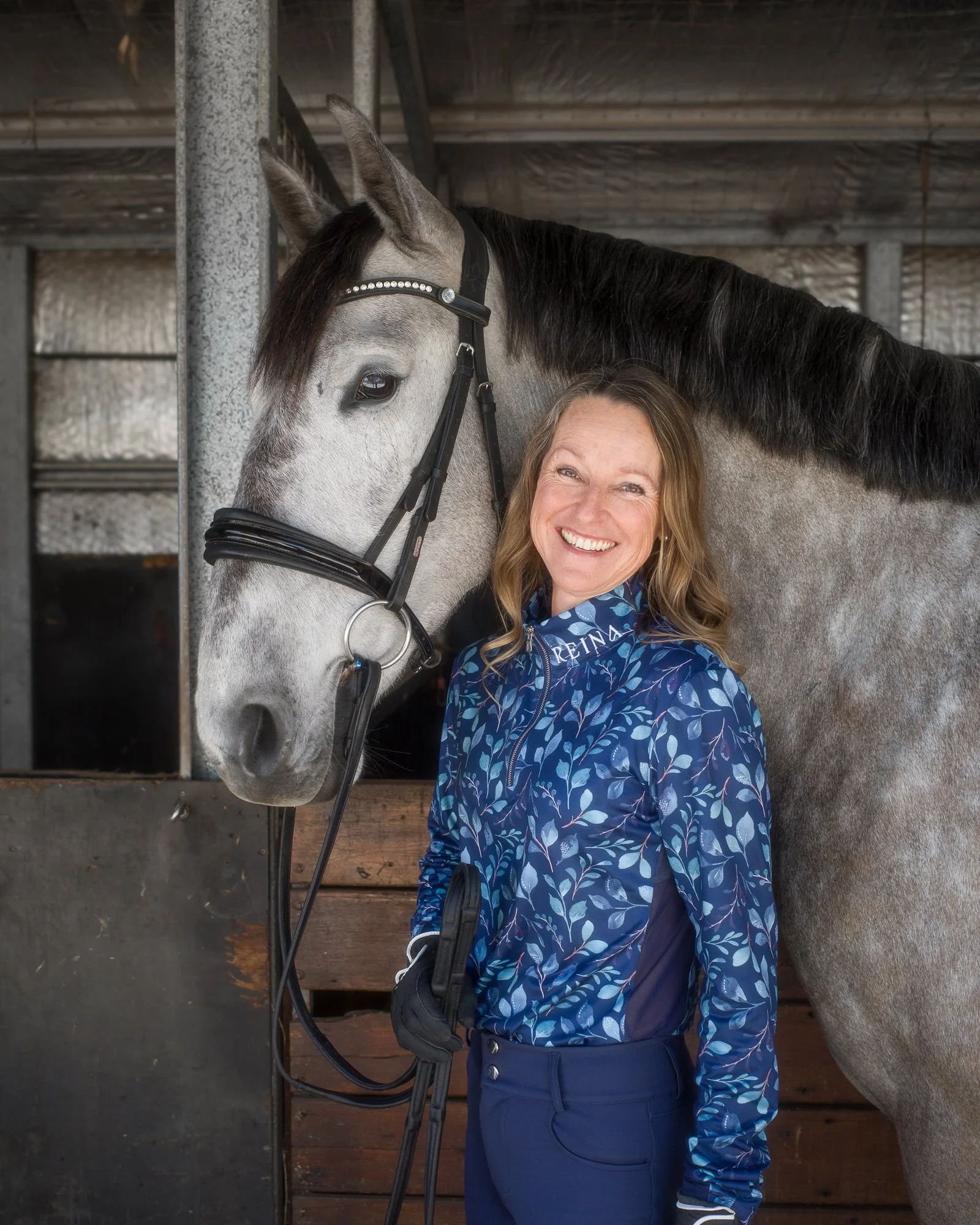 A woman with light brown hair, smiling, standing next to a gray and white horse with a black mane. The woman is wearing a blue patterned jacket with the word 'REINA' visible and dark blue pants. The horse is wearing a bridle with decorative stones. They are in a stable with wooden and metal walls in the background.