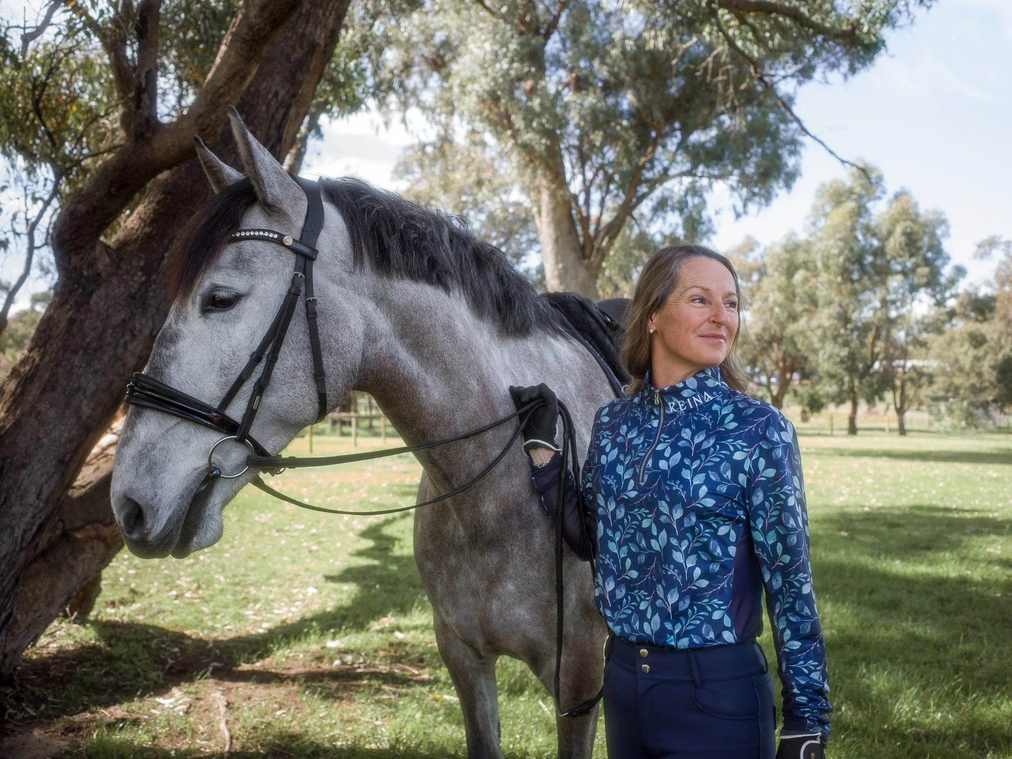 A woman standing next to a gray horse under a tree in a park-like setting with trees and grass, sunny weather, and a partly clouded sky.