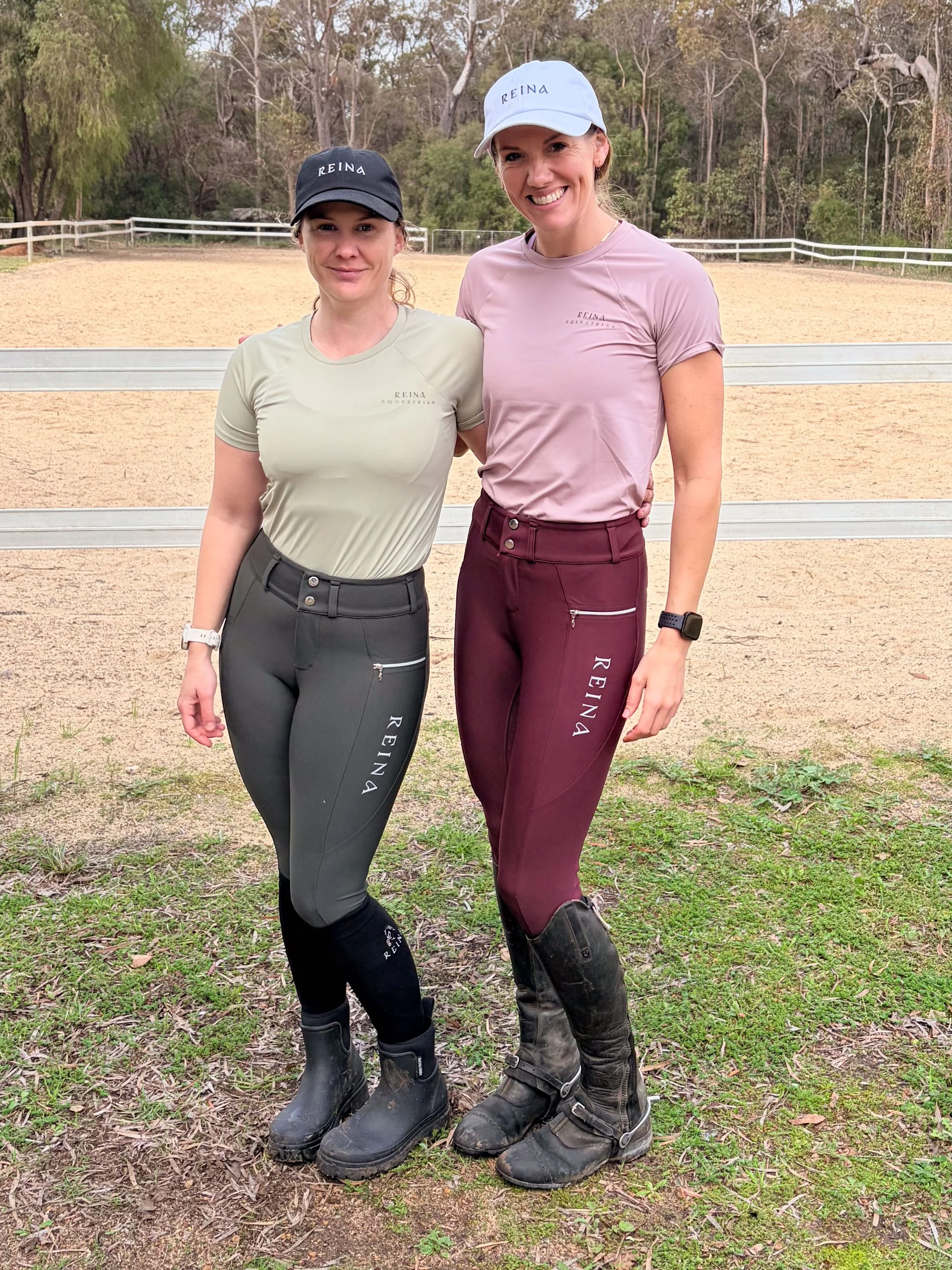 Two women standing outdoors on a riding arena with trees and a wooden fence in the background, both wearing riding gear including boots, riding pants, and hats with 'REINA' logo.