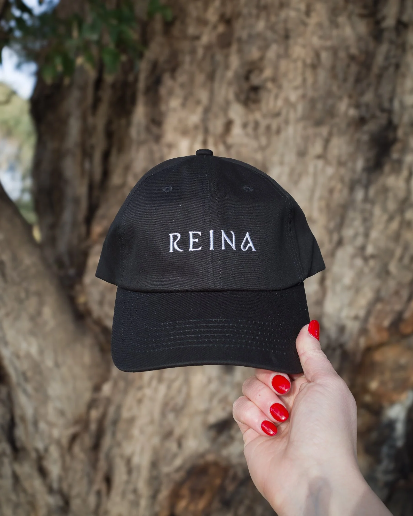 A person with red painted nails holding a black baseball cap with the embroidered text 'REINA' on it, in front of a large tree trunk.
