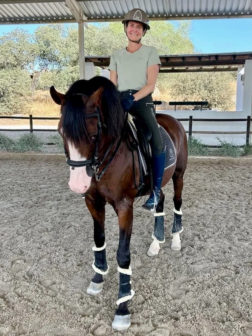 A woman wearing riding gear and a helmet sits on a brown horse with a white face inside an indoor horse riding arena. The horse has protective boots on all four legs.