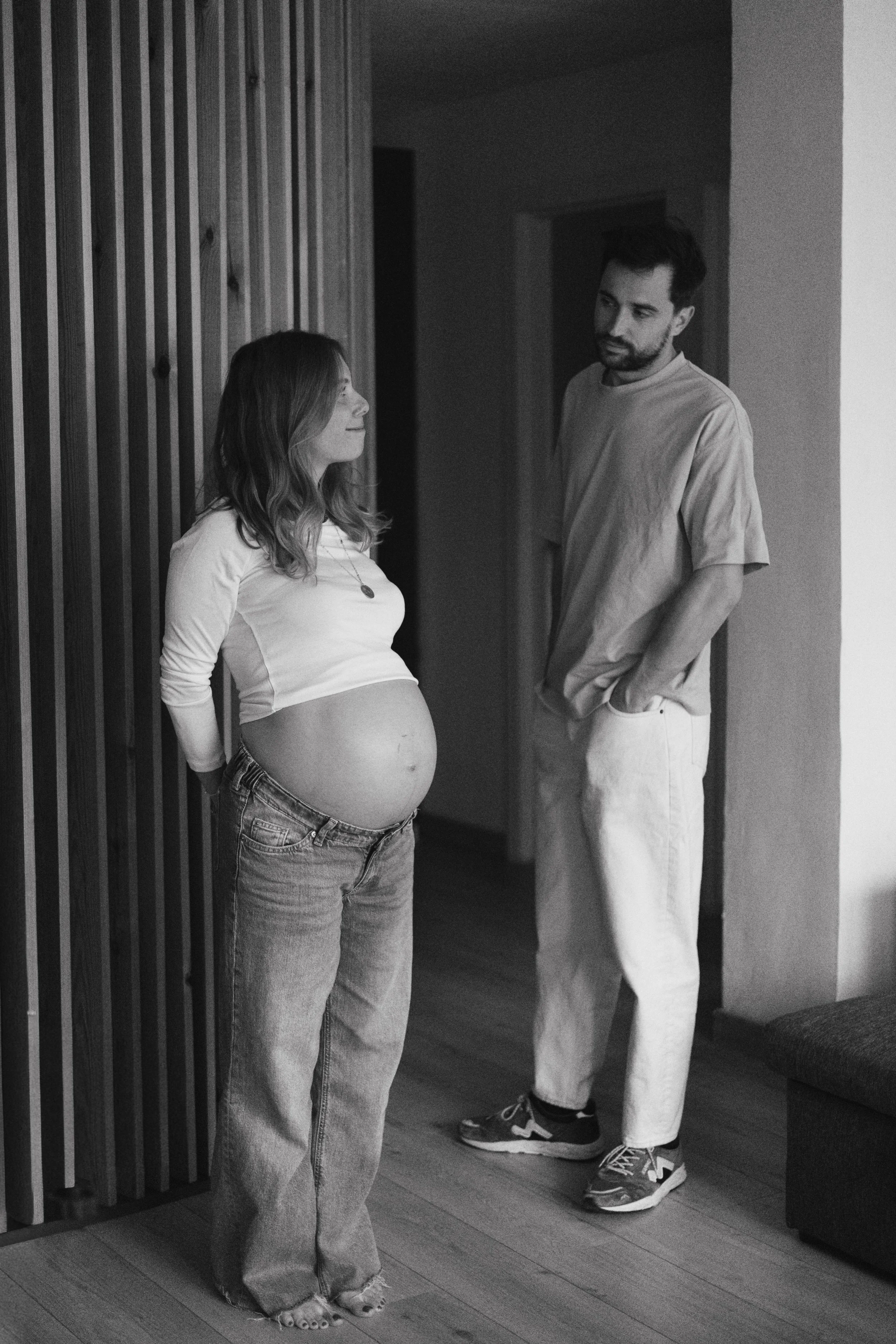 Une femme enceinte assise contre un mur en bois, parlant à un homme debout à côté d'elle dans un intérieur