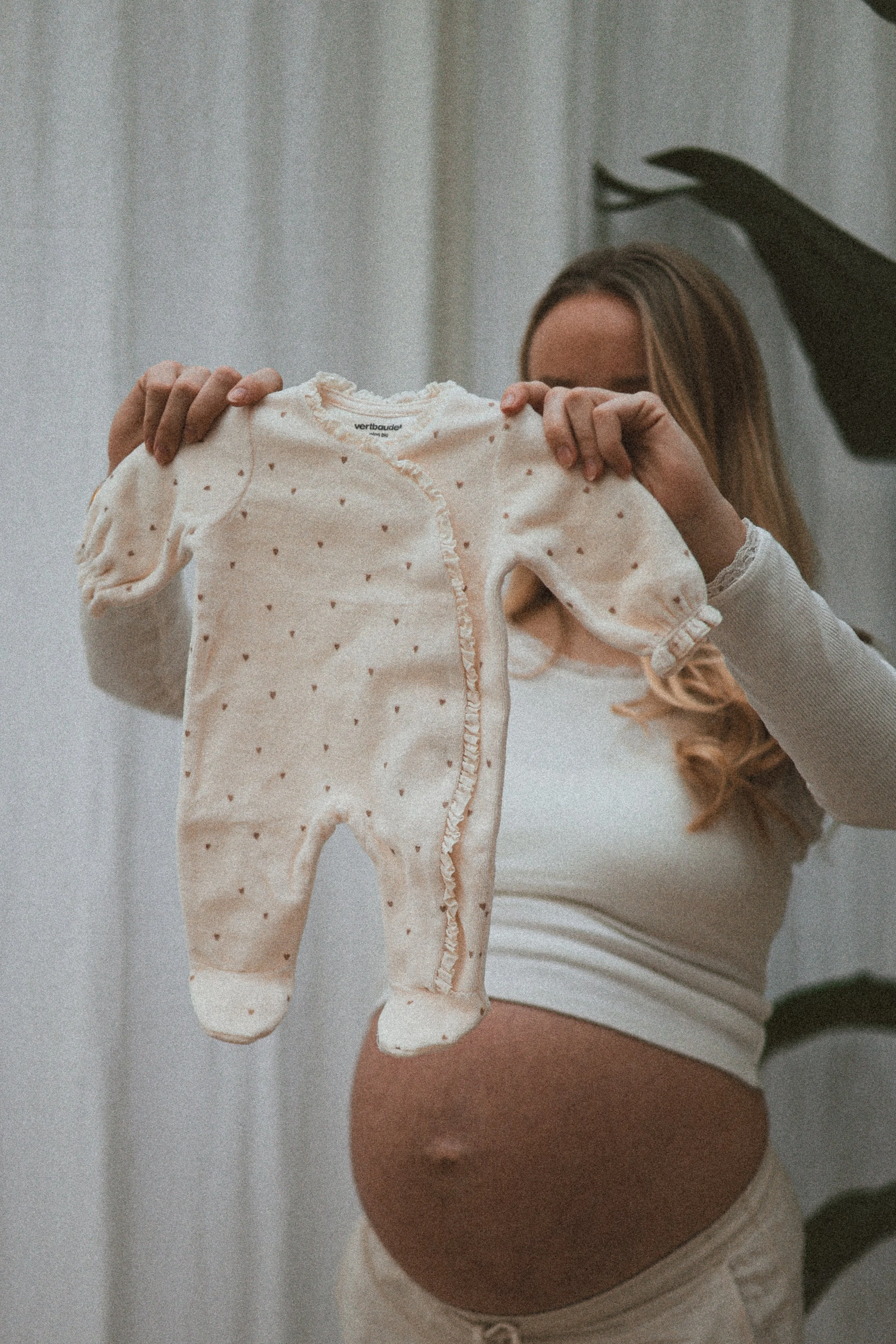 Une femme enceinte tient un pyjama pour bébé en velours beige à pois marron, devant un miroir, dans une pièce lumineuse.