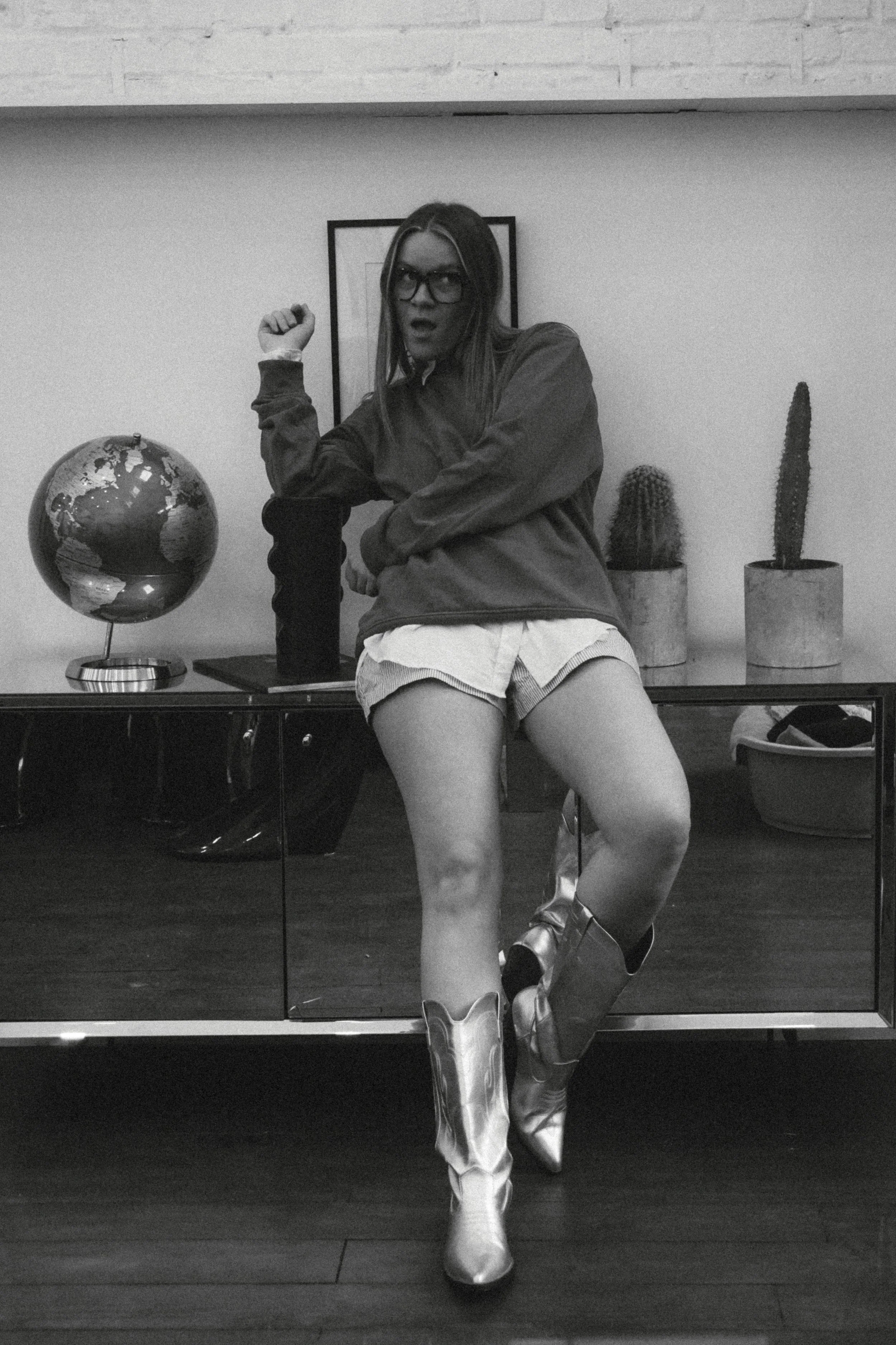 Une femme portant des bottes de cowboy hautes, une robe courte et une veste, pose devant un miroir. Elle porte des lunettes et a les cheveux longs. Il y a un globe terrestre et deux cactus en pots sur une étagère derrière elle.