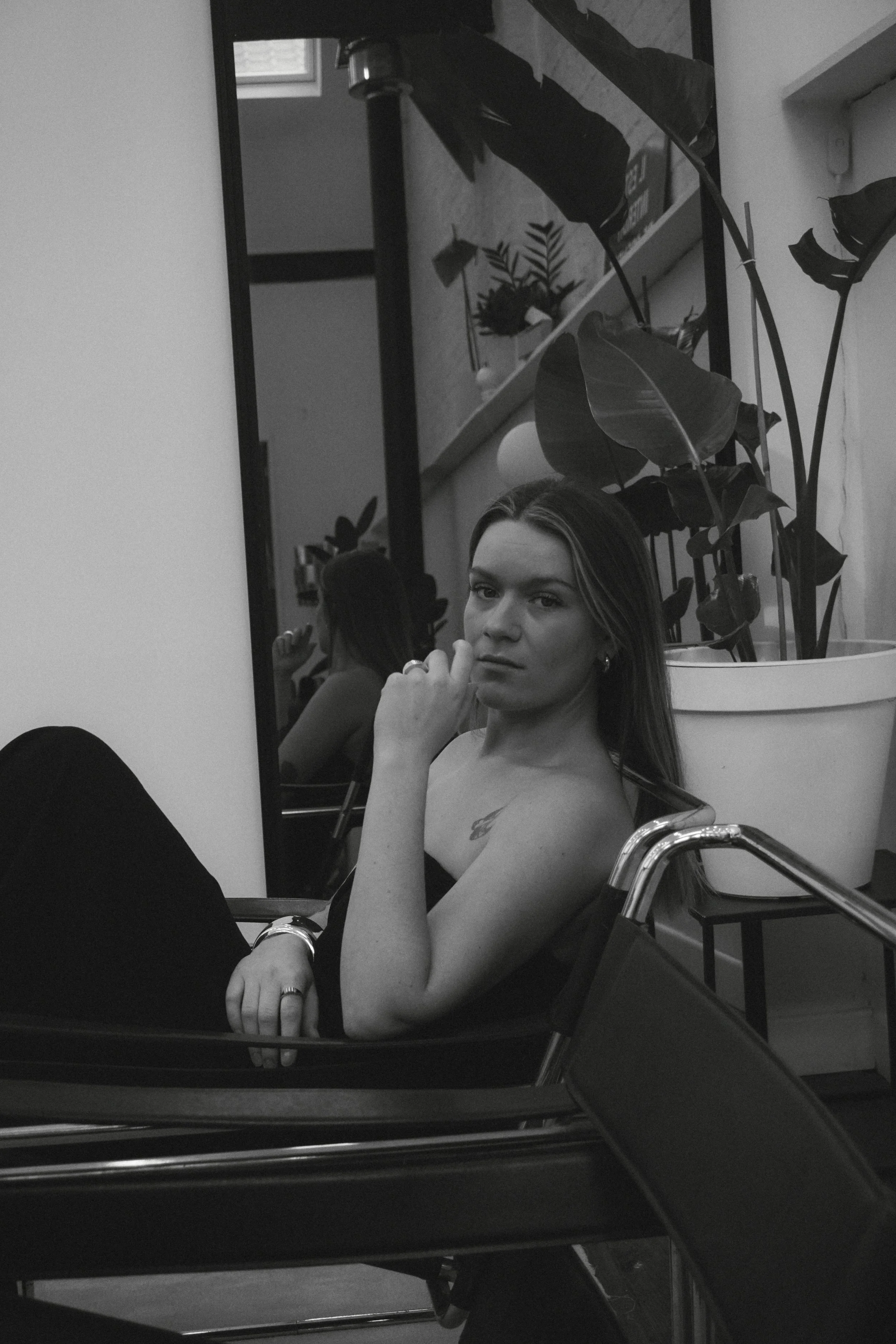 Une femme regardant la caméra dans un miroir, assise à une table, avec une plante en arrière-plan.