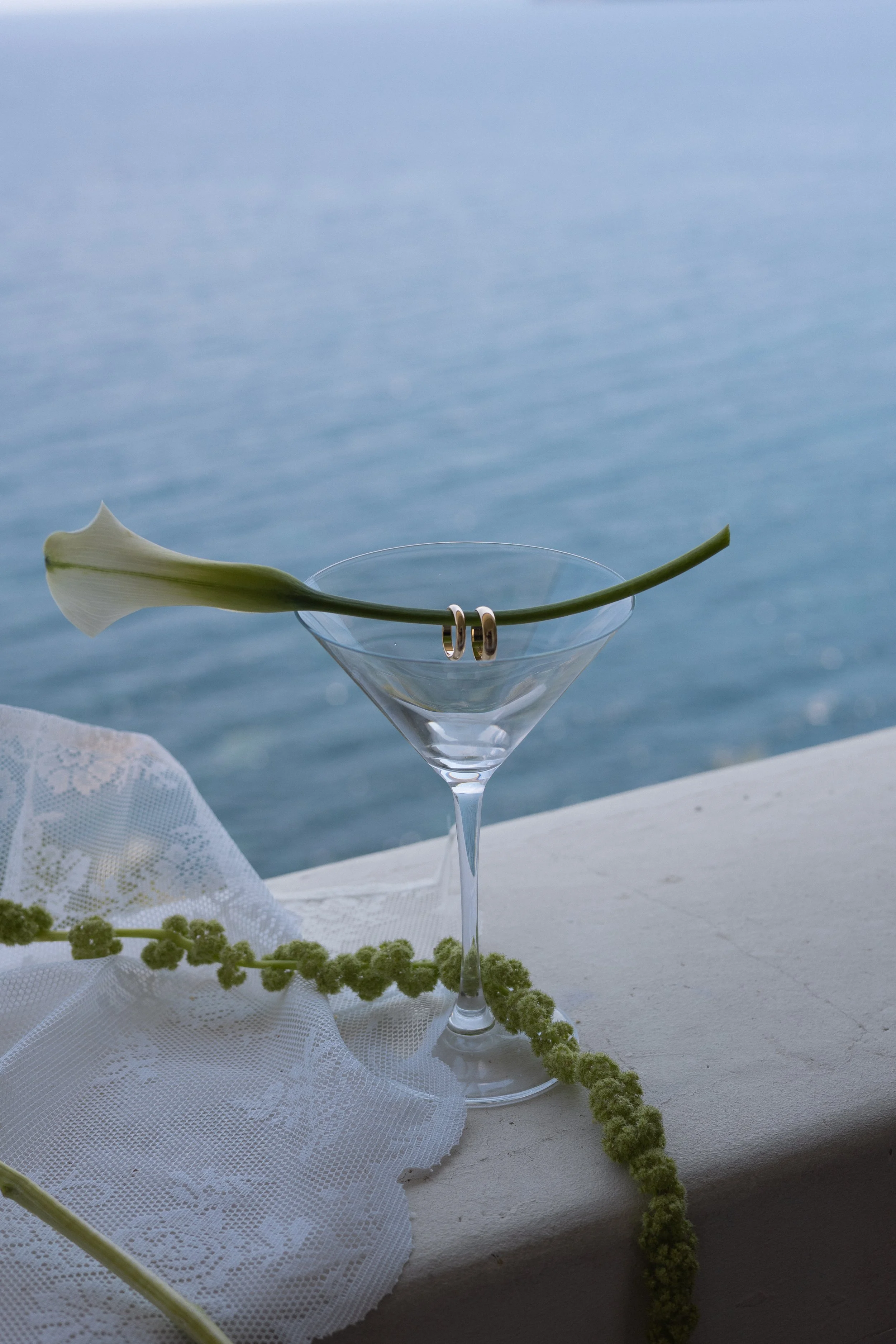 Une coupe à cocktail vide avec une calla lily blanche posée dessus, un fond bleu et une toile blanche avec des fleurs vertes en premier plan.