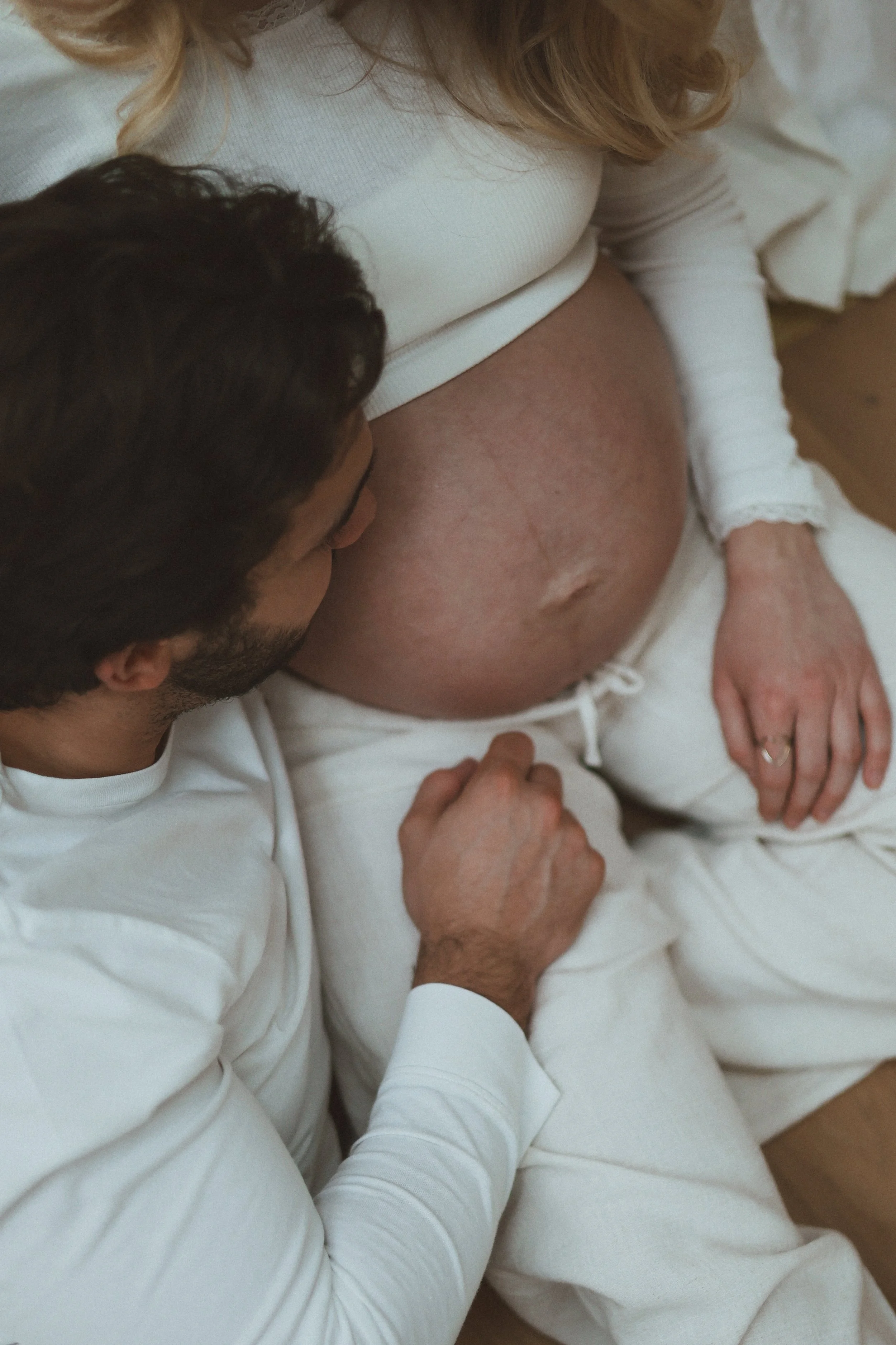Une femme enceinte et un homme dans une pièce, partageant un moment intime, la femme porte un haut blanc et des vêtements confortables