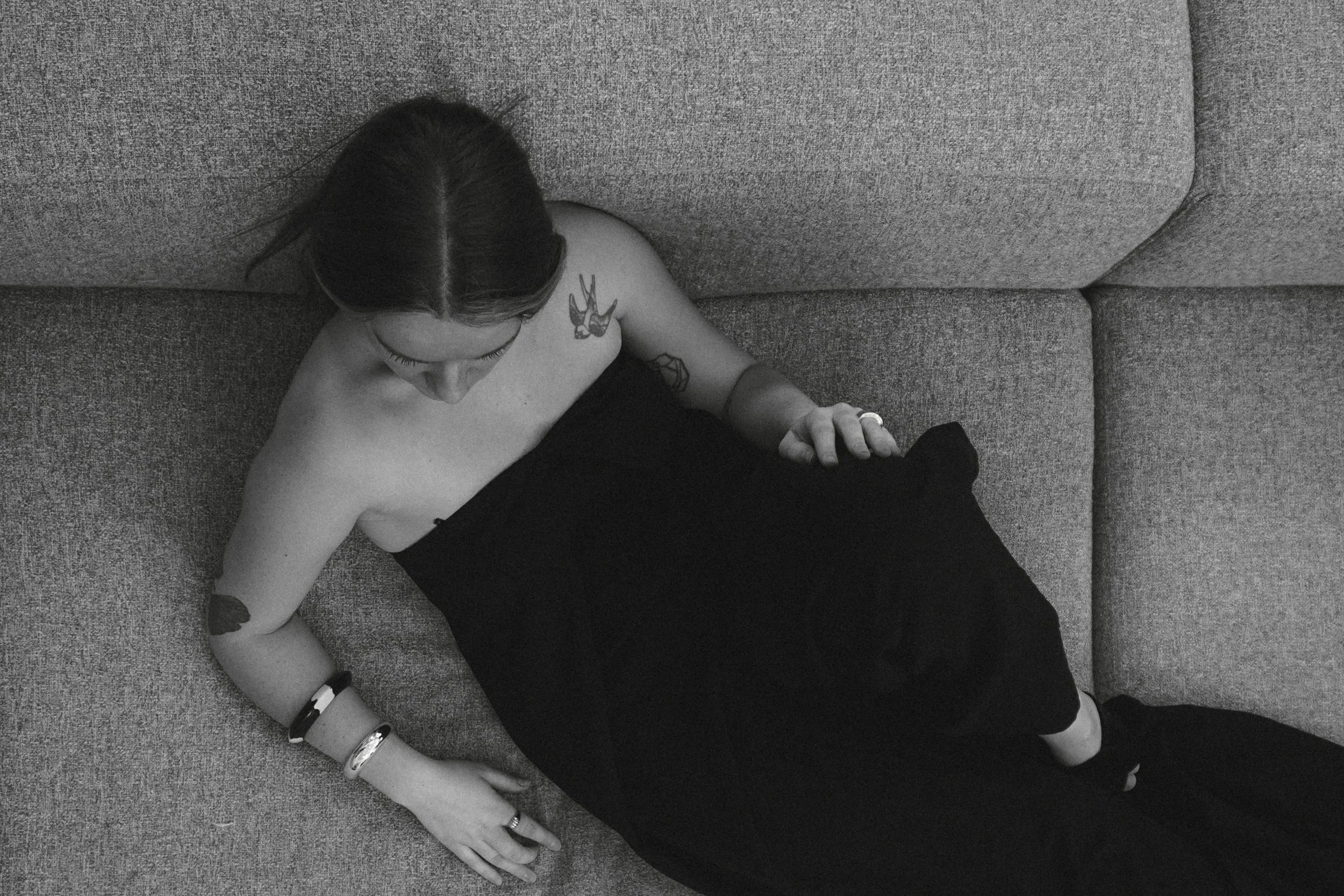Une femme allongée sur un canapé, vue de haut, portant un haut noir et plusieurs bracelets. Ses tatouages sont visibles sur l'épaule et le bras, ses yeux sont fermés.