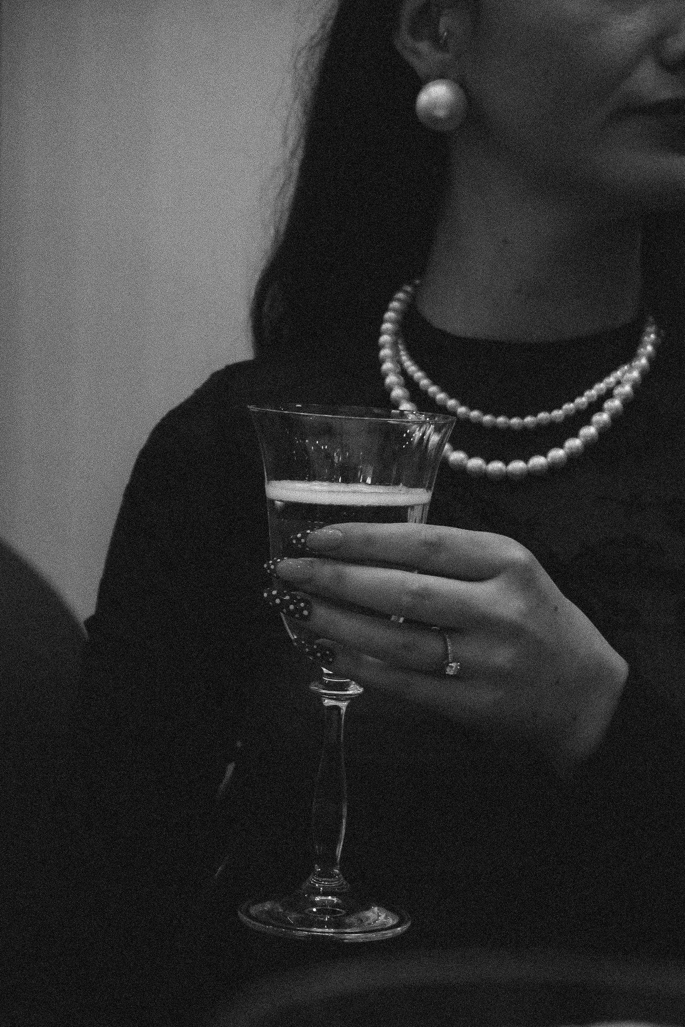 Une femme tient un verre à vin dans une main et porte des bijoux, notamment des perles et une paire de boucles d'oreilles, en noir et blanc.