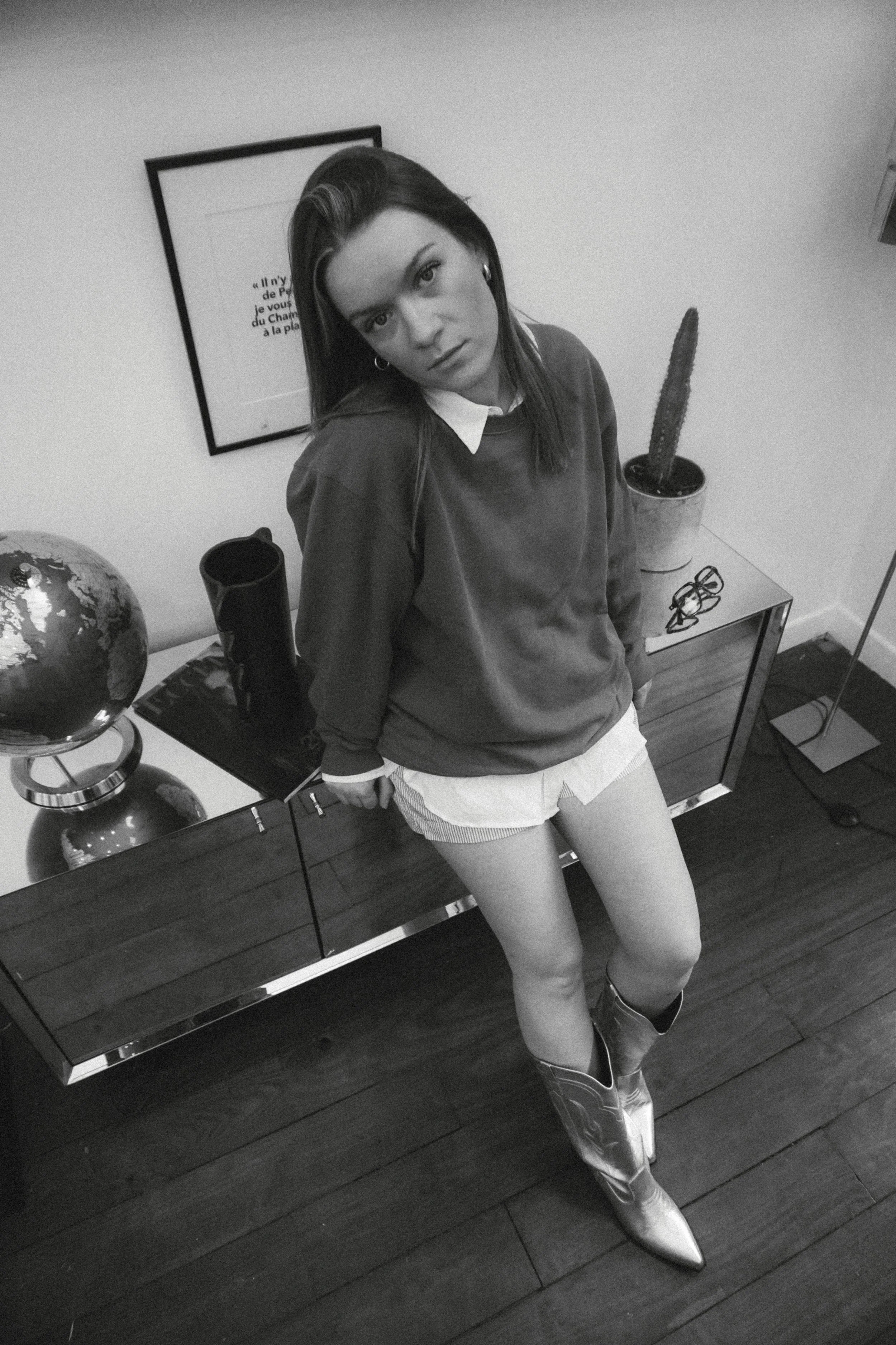 Une femme en tenue à la mode avec des bottes en cuir, posant dans une pièce avec un décor minimaliste comprenant une table en bois, un globe terrestre, une plante cactus, une paire de lunettes et un tableau au mur. C'est une photographie en noir et blanc.