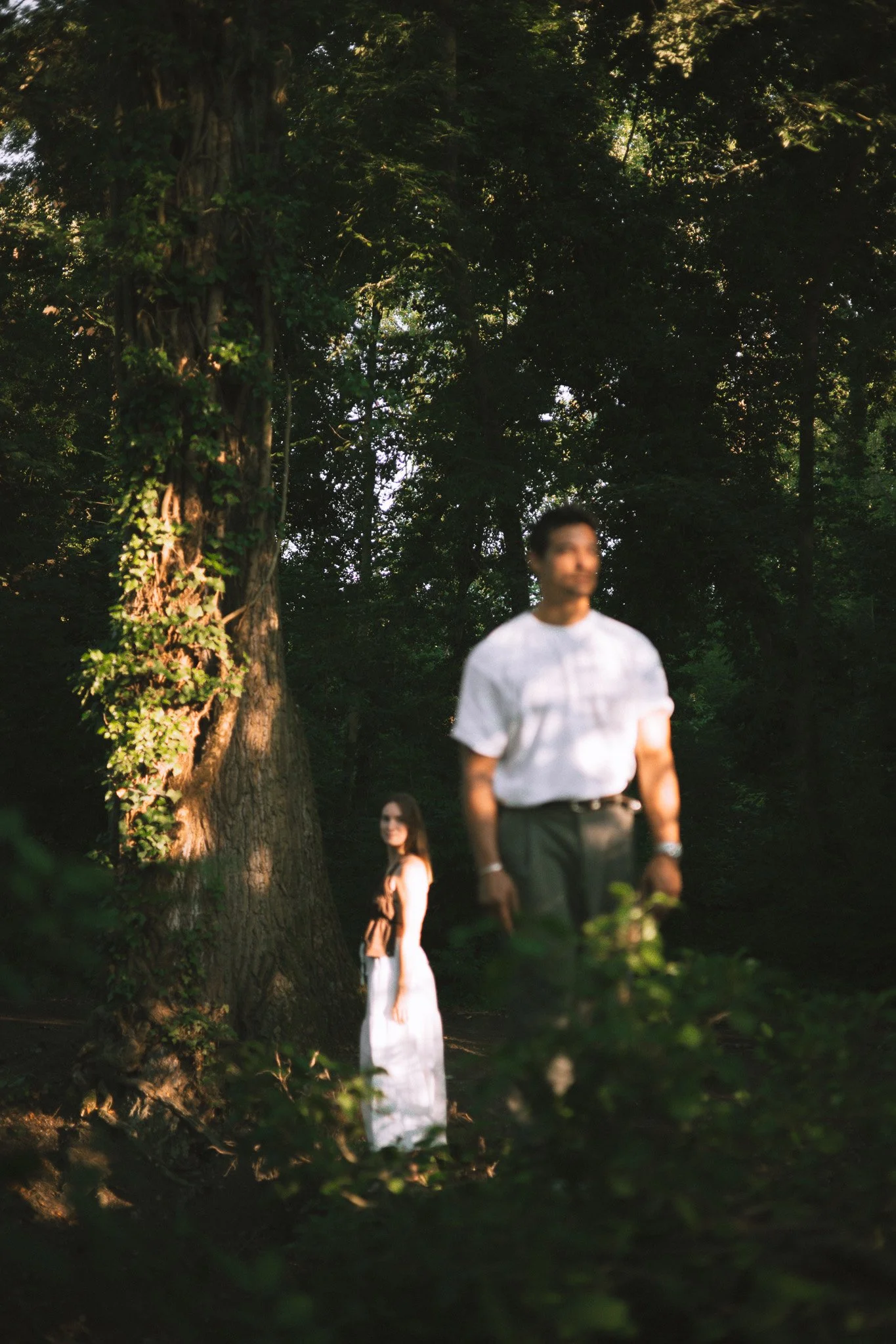 Une personne portant une chemise blanche et un pantalon gris, et une femme en robe blanche, se tenant à l'extérieur près d'un arbre, dans un environnement boisé avec de la lumière naturelle filtrée à travers les arbres.