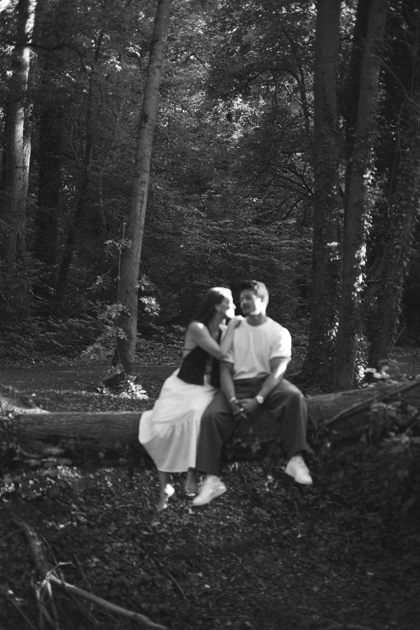 Deux personnes, un homme et une femme, assises sur un tronc d'arbre dans une forêt. La femme porte une robe blanche et l'homme une chemise blanche, ils semblent discutent ou partagent un moment intime. La scène est en noir et blanc.