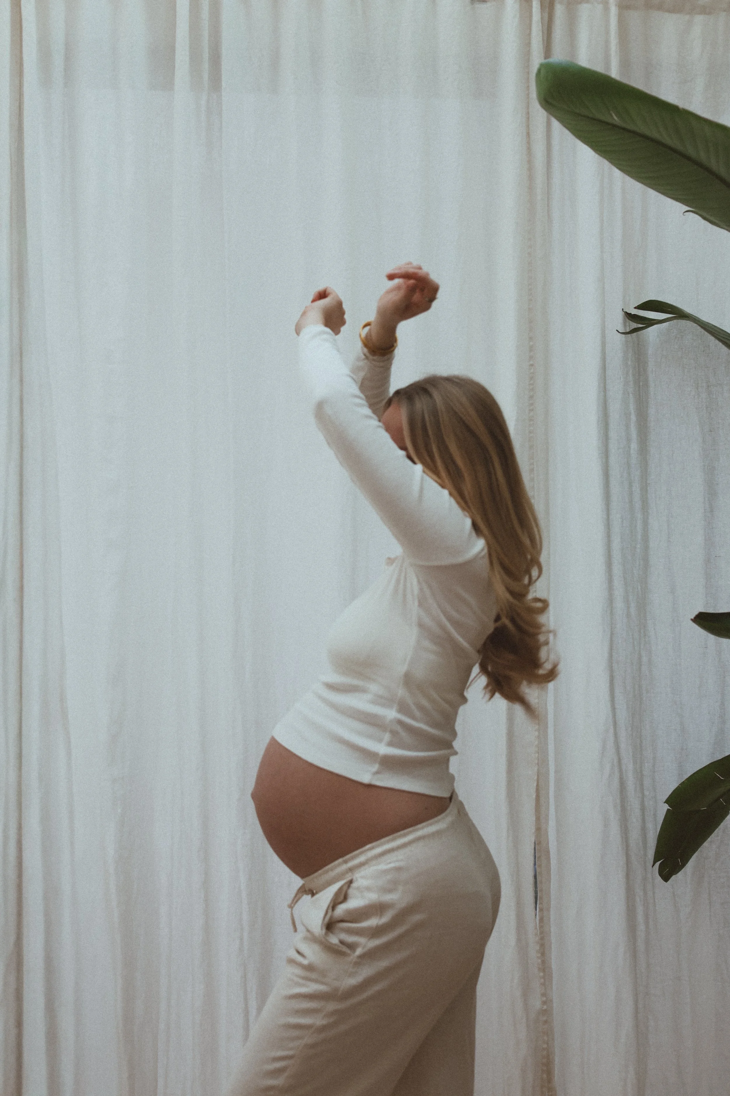 Femme enceinte posant en lingerie blanche dans une pièce avec des rideaux clairs et un grand feuillage vert à droite.