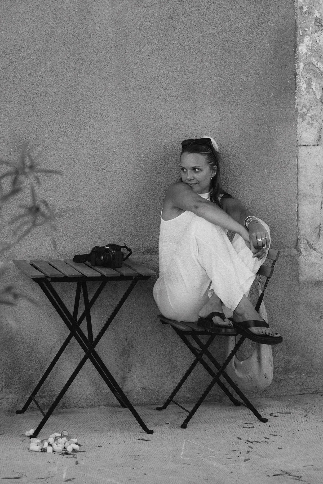 Une femme assise sur une chaise pliante, portant une robe blanche, des lunettes sur la tête, et regardant sur le côté. Elle a des bracelets à son poignet et une bague sur son doigt. Il y a un appareil photo sur une table en face d'elle. Au sol, il y a un tas de balles de golf ou de plastique blanc, ainsi que des débris et une cuillère. L'arrière-plan est un mur en pierre et en plâtre.