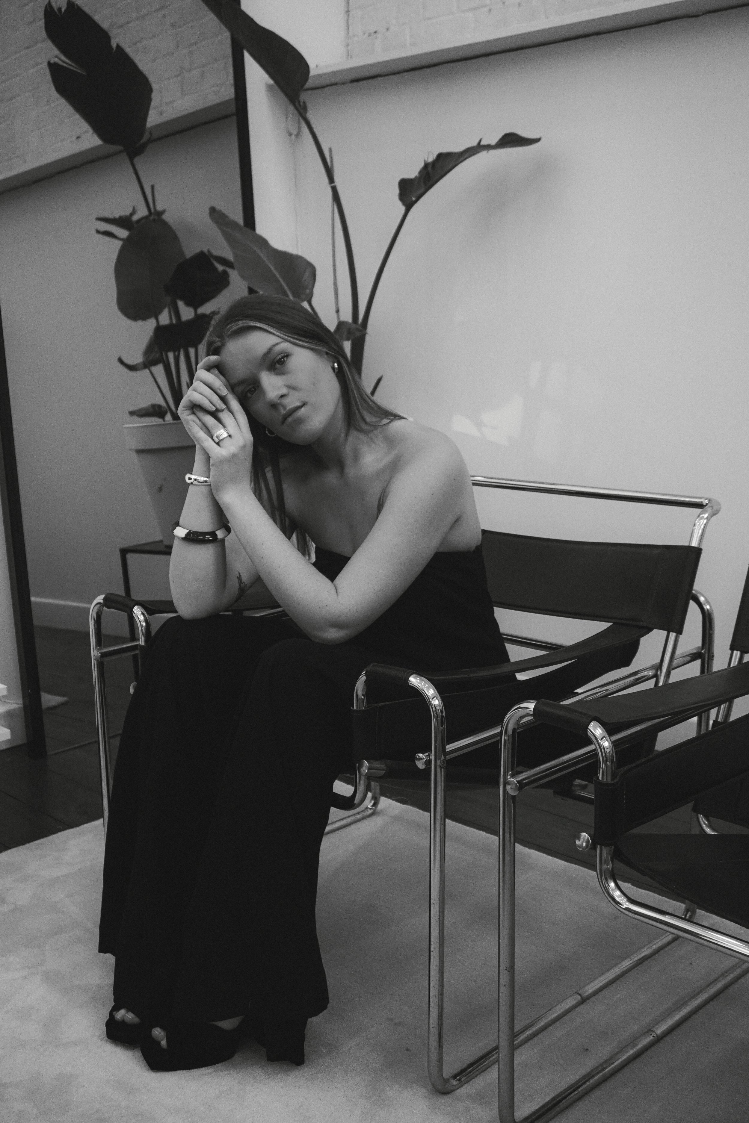 Une femme assise dans une salle, portant une robe longue noire, les cheveux longs et droits, avec des bracelets et une bague, posant avec sa main contre sa tête, derrière une plante verte en pot et des chaises en métal.