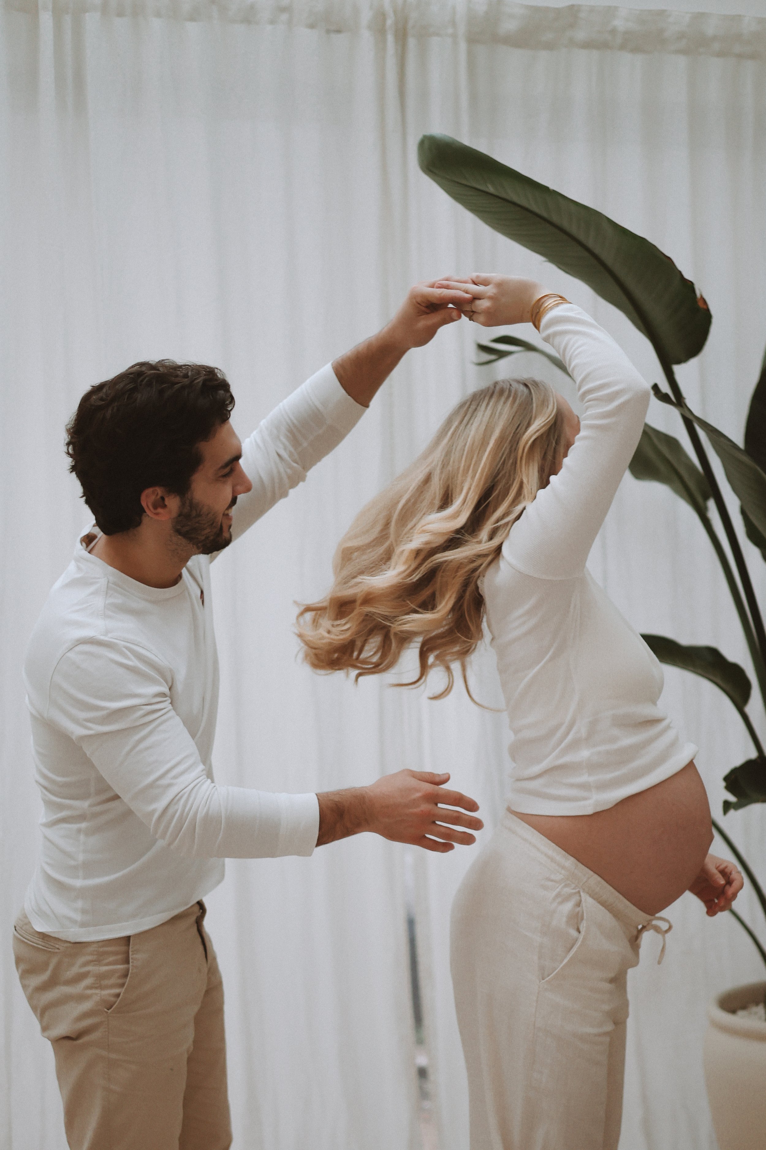 Une femme enceinte avec des cheveux longs et bouclés posant avec un homme dans un décor intérieur avec des rideaux blancs et des plantes vertes. L'homme et la femme semblent danser ou faire une séance de photographie artistique, la femme portant un haut blanc et un pantalon beige, et l'homme portant une chemise blanche et un pantalon beige.