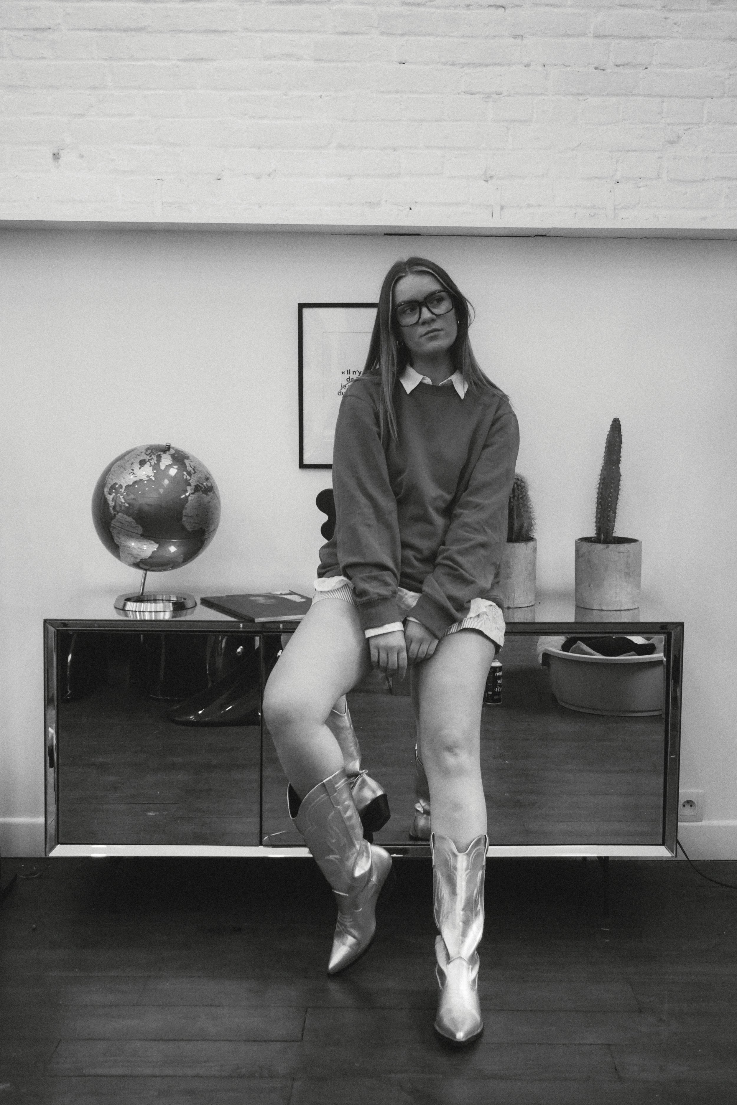 Une jeune femme portant des bottes en cuir clair, une jupe courte, un sweat-shirt gris, une chemise blanche en dessous et des lunettes, pose devant un miroir dans une pièce décorée avec un globe terrestre, deux cactus en pot et un cadre avec une citation, dans une ambiance contemporaine.