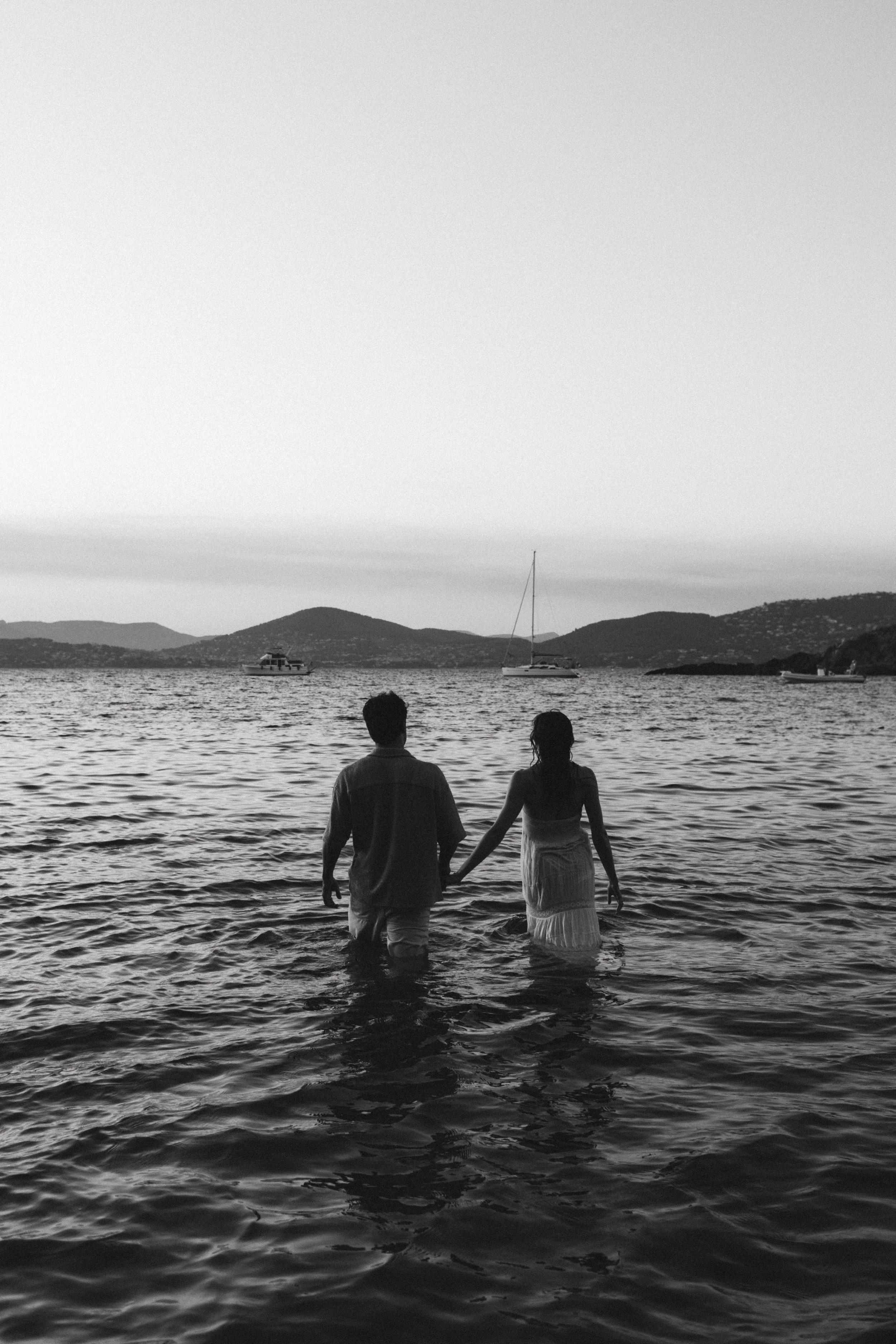 Deux personnes, un homme et une femme, tenant la main, marchent dans l'eau de mer près de la plage, avec des bateaux et des îles en arrière-plan, en fin de journée ou au coucher du soleil.