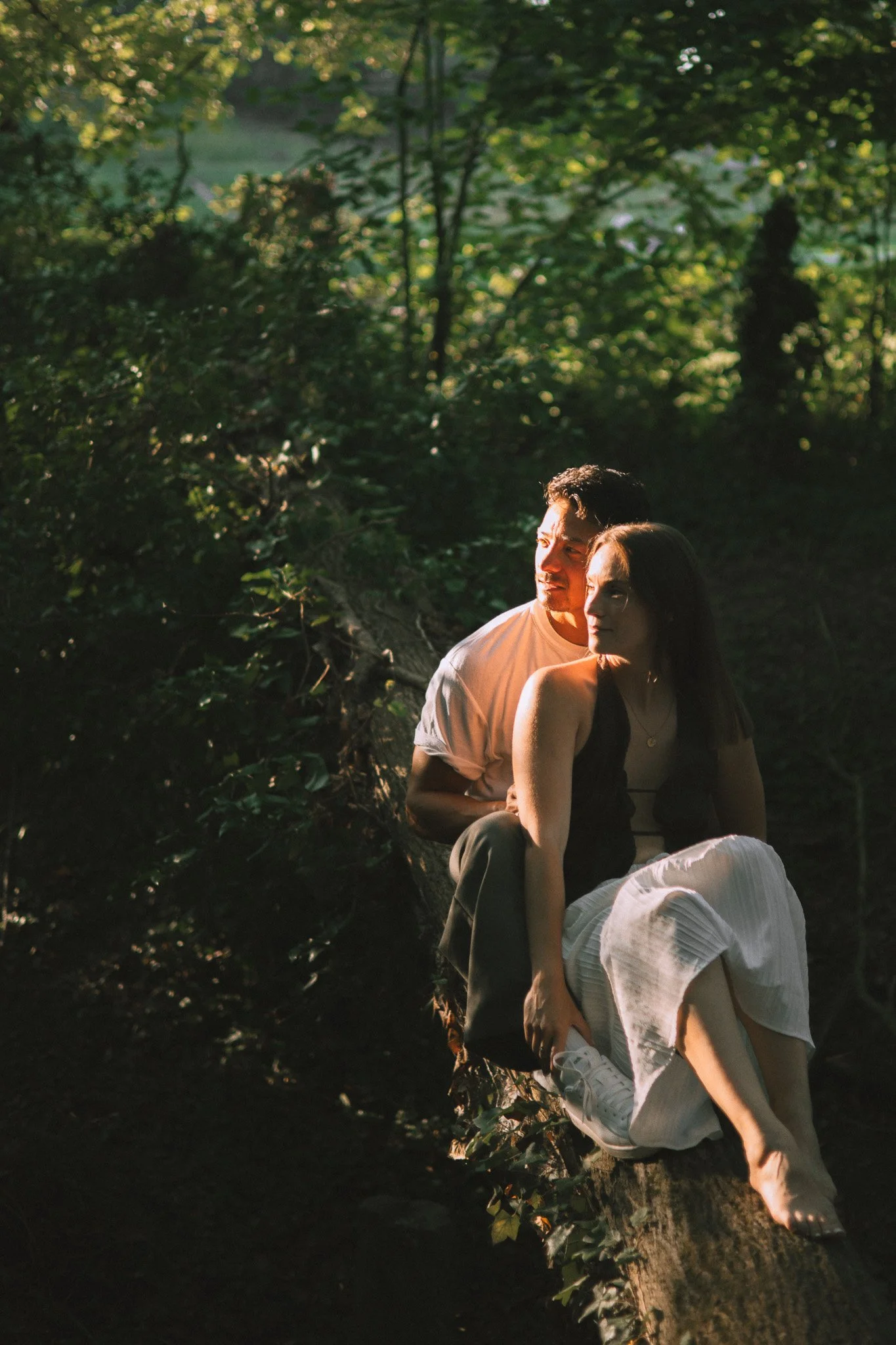 Deux jeunes adultes assis sur un arbre dans une forêt verdoyante éclairée par le soleil, l'homme regarde au loin tandis que la femme regarde le sol.