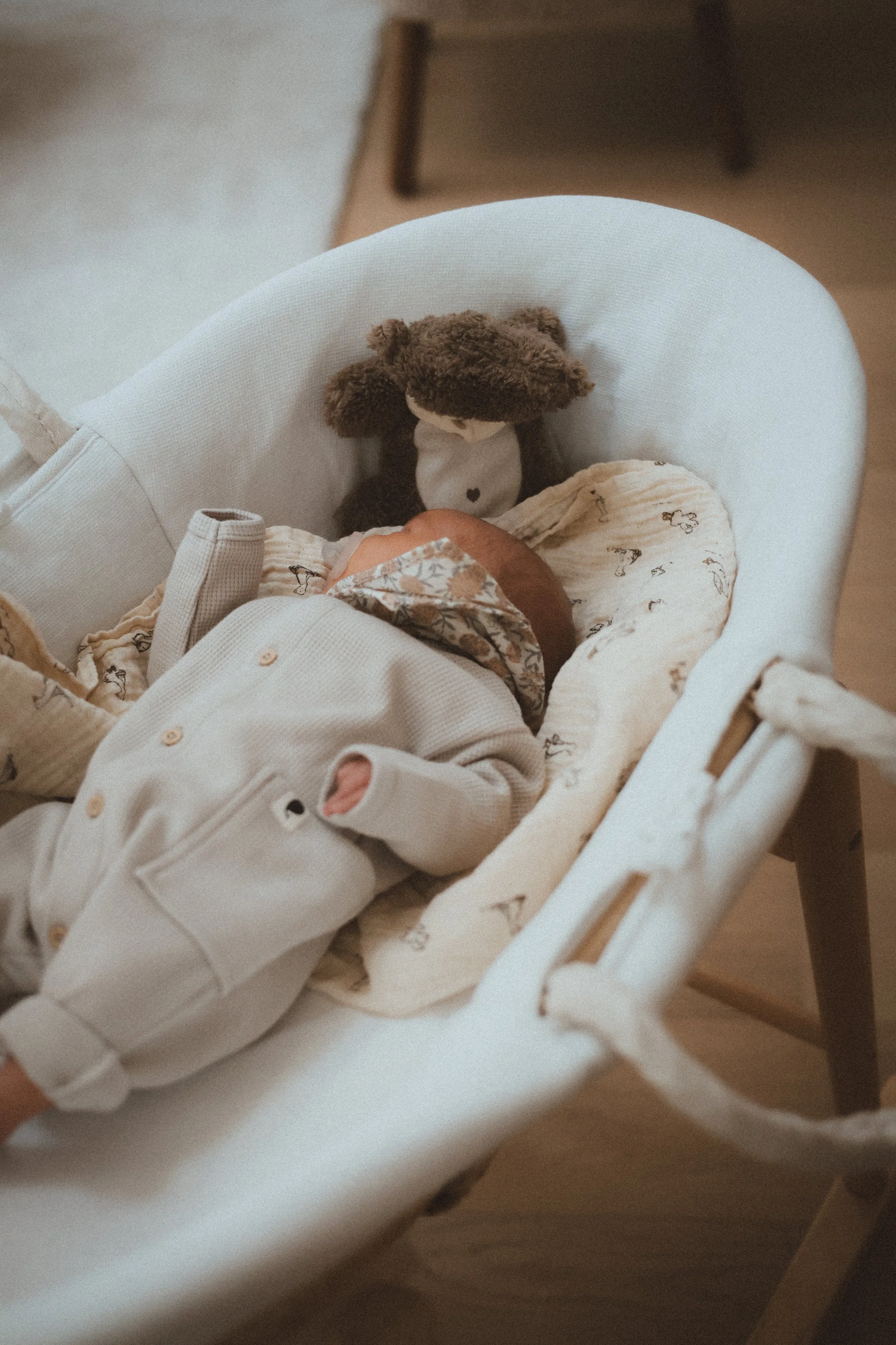 Un bébé endormi dans un couffin en tissu blanc, vêtu d'une tenue beige avec des petits motifs, avec une peluche de singe posée devant lui.