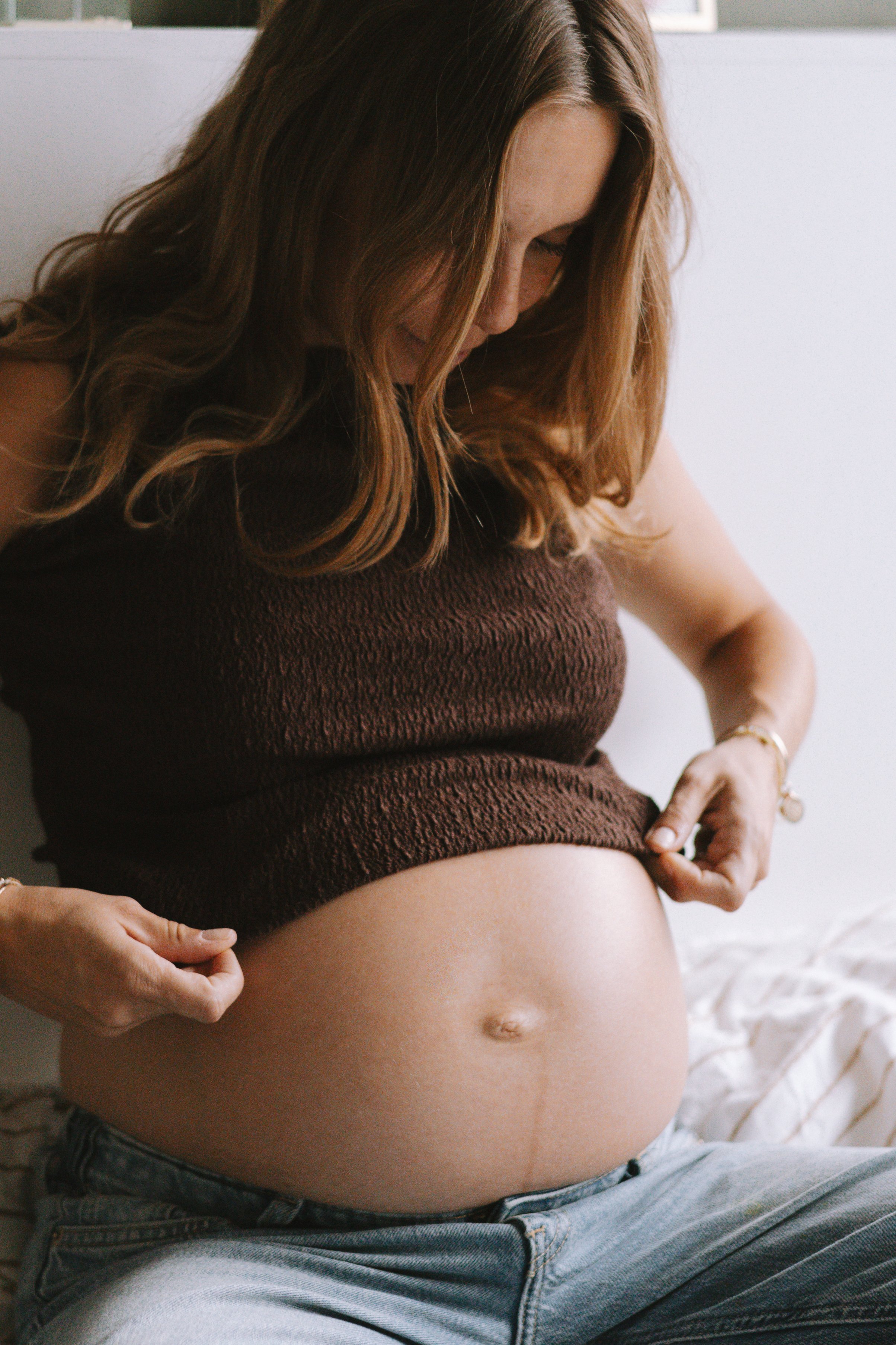 Une femme enceinte en pantalon en jean et haut marron, assise ou allongée, avec son ventre nu visible. Elle tient ses mains sur son ventre, dans un environnement intérieur.