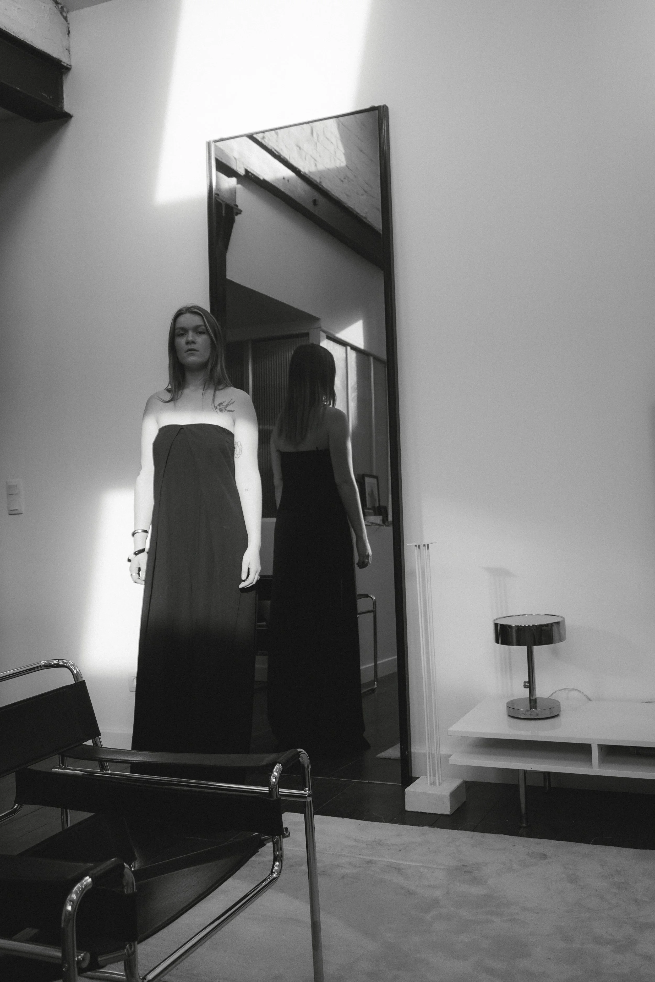 Une femme avec des cheveux longs porte une robe longue dans une pièce moderne, vue à travers un miroir rectangular. La pièce a des meubles noirs, une table blanche avec une lampe argentée et un fauteuil noir.