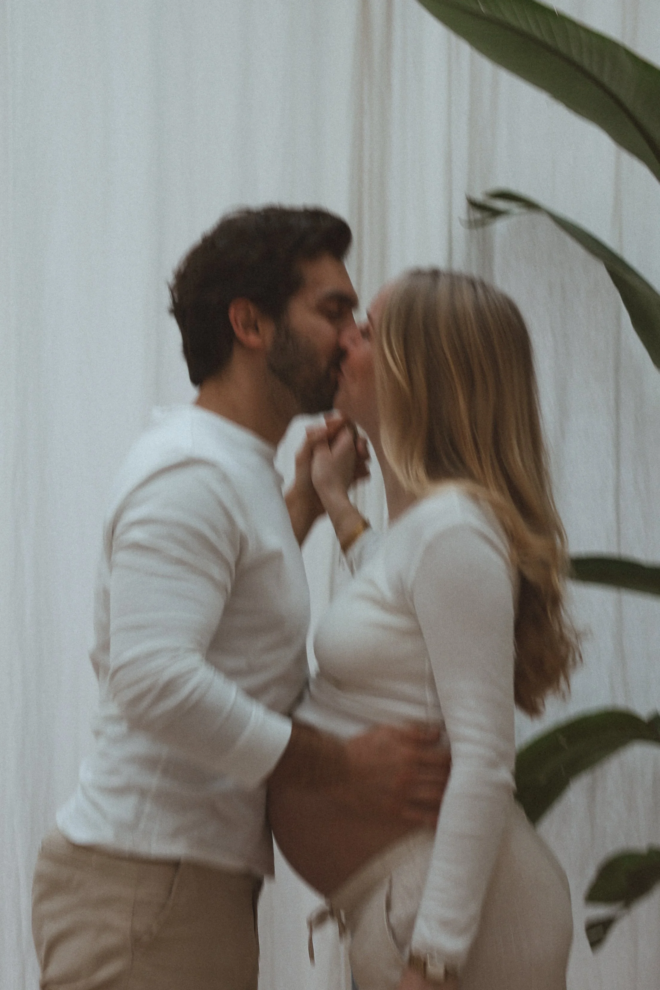Un couple s'embrasse et se tient près d'un mur blanc, avec une grande plante verte à droite.