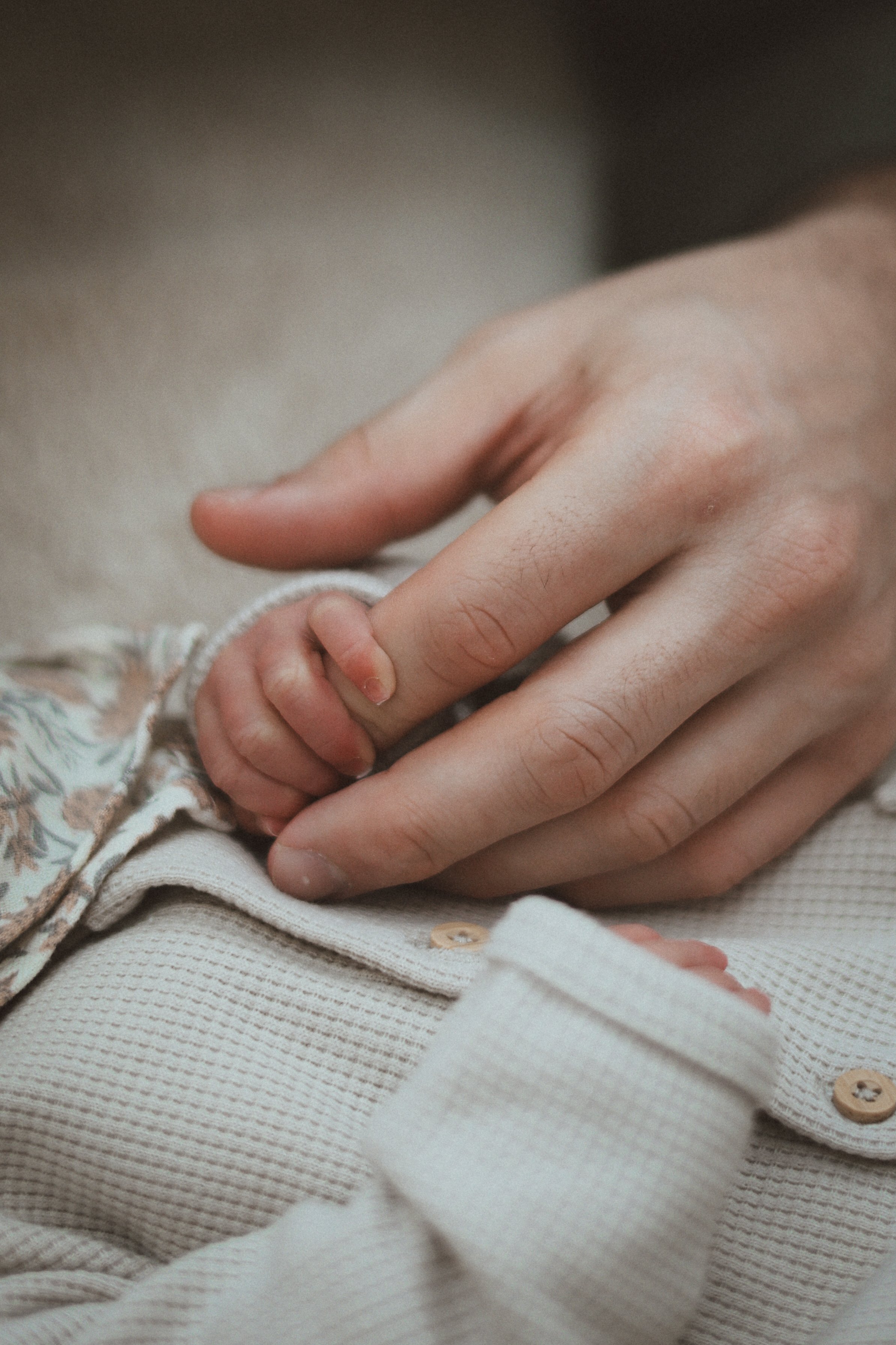Une main d'adulte tenant doucement une main d'enfant, reposant sur une surface en tissu beige, vêtue d'une chemise à manches longues à motif floral et d'un pull beige.